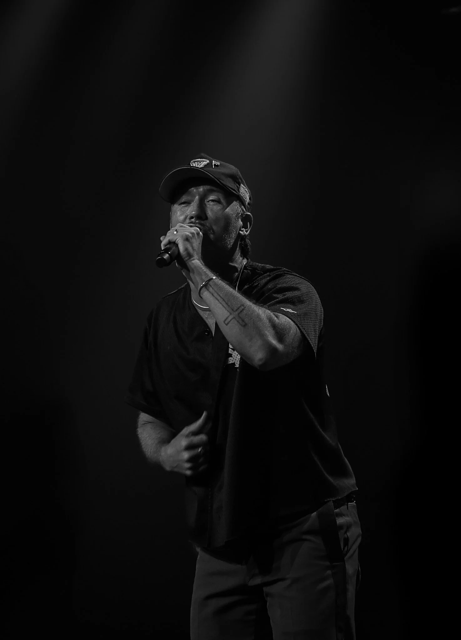 Black and white photo of a man singing into a microphone on stage, wearing a cap, with tattoos on his arm, and a serious expression.