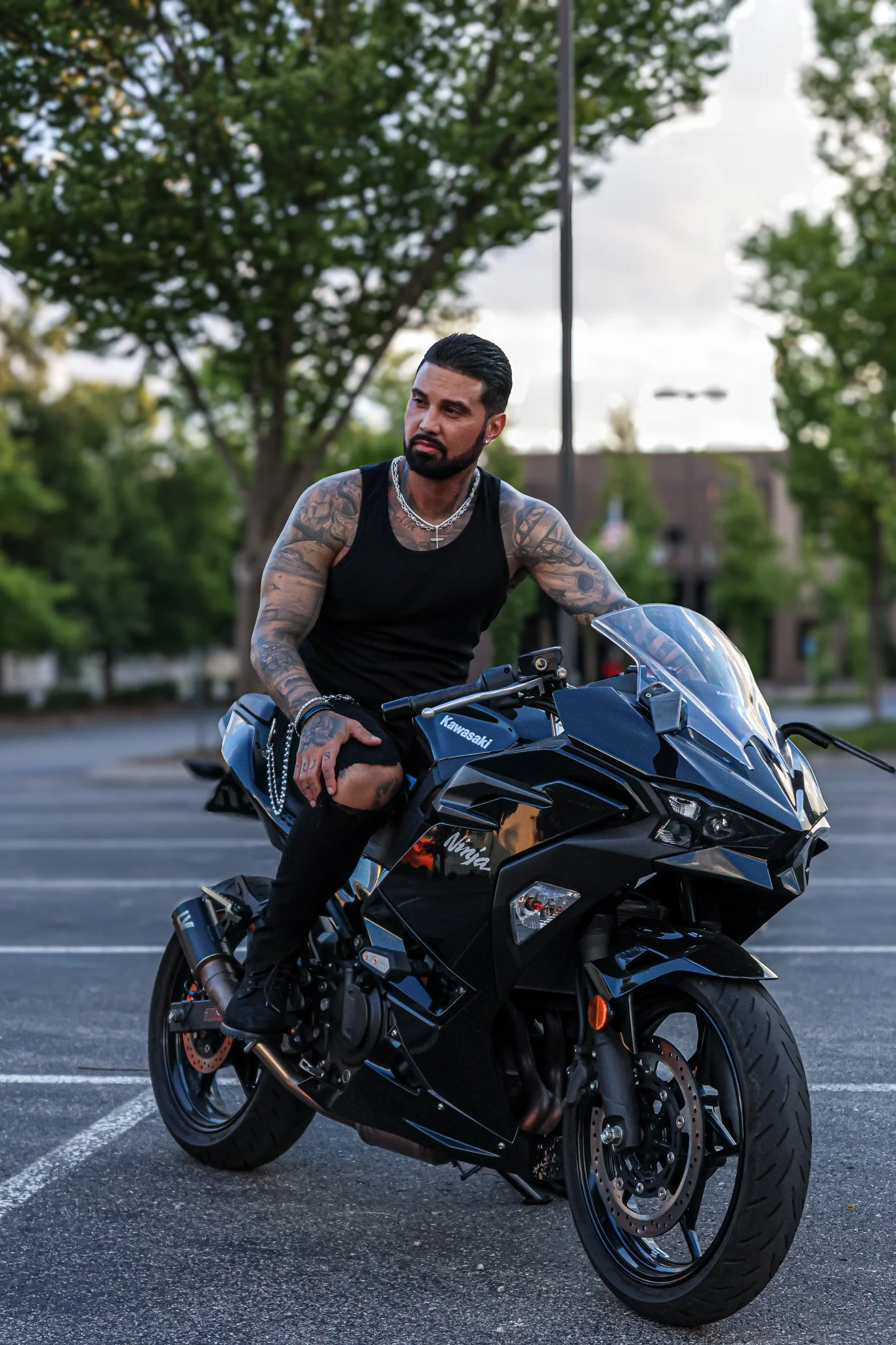 A man with tattoos wearing a black tank top and ripped jeans sits on a black Kawasaki Ninja motorcycle in an empty parking lot with trees and buildings in the background.
