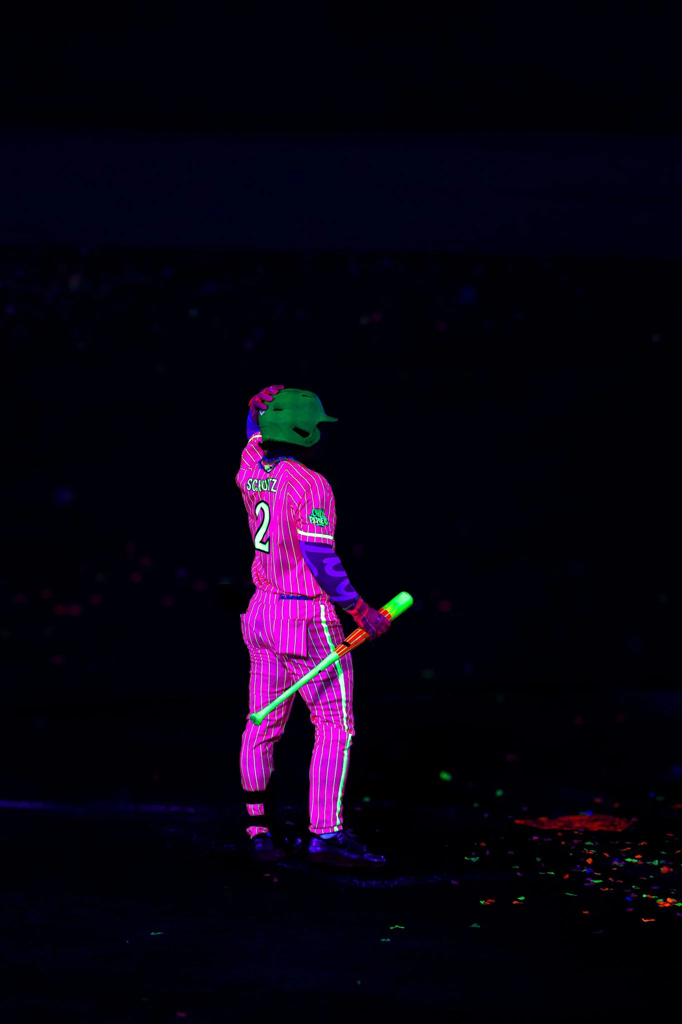 Person wearing a glow-in-the-dark sports uniform, helmet, and gloves holding a glowing baton, standing on a dark surface with scattered glowing confetti.