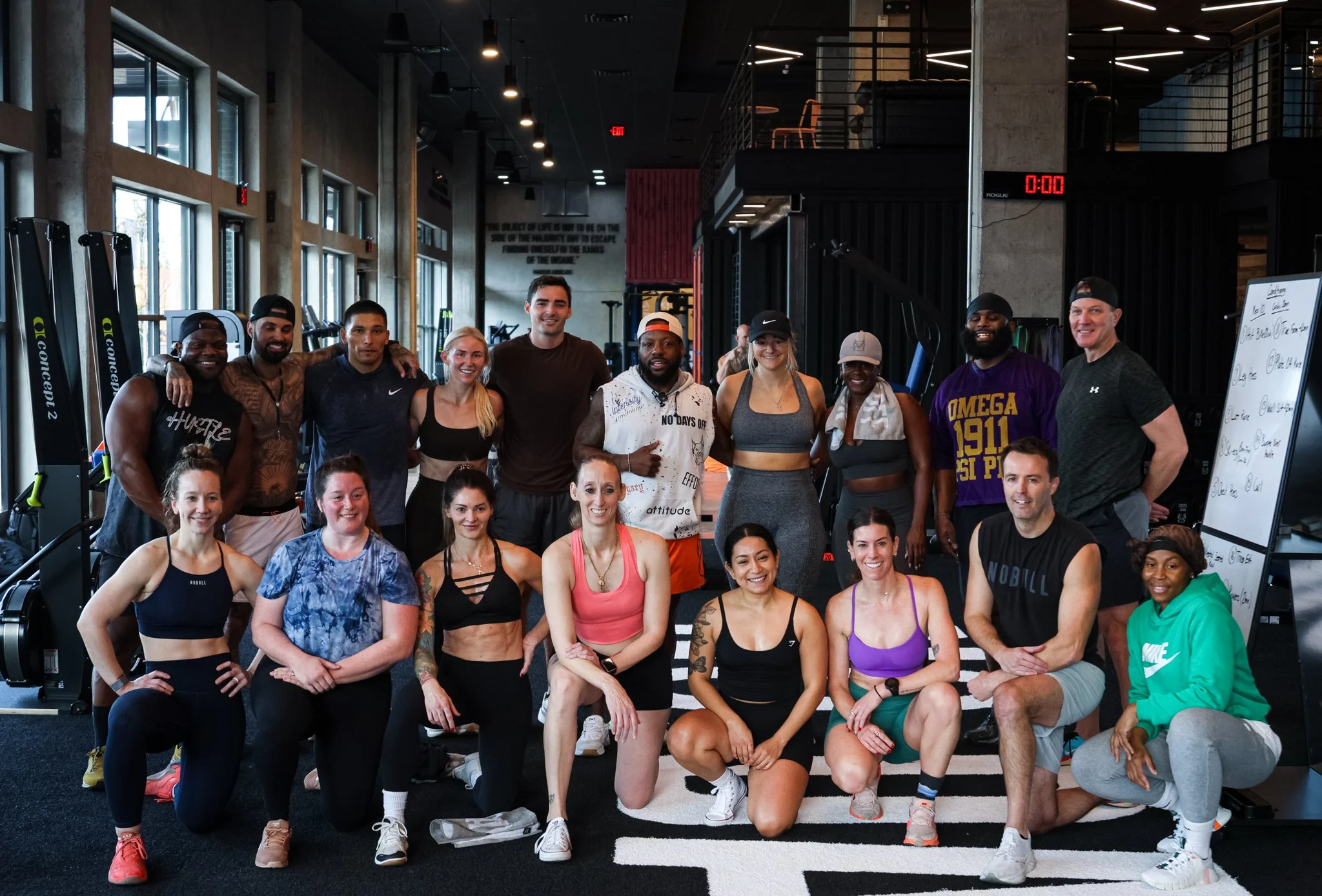 Group of fit individuals in workout attire posing together inside a modern gym with exercise equipment and large windows.