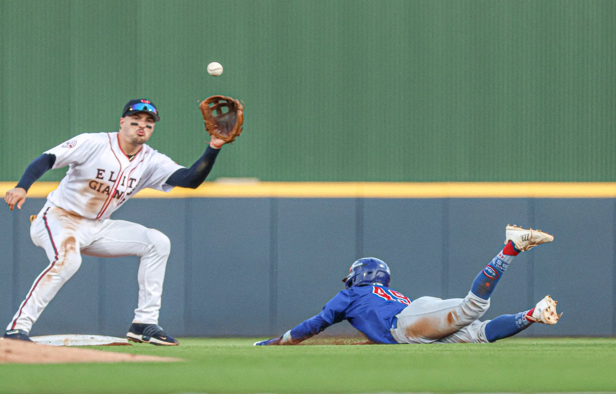 A baseball player from the white team, labeled 'ELITE GIANTS,' catches a baseball with his glove near second base while a player from the blue team, labeled with the number 45, slides into the base.