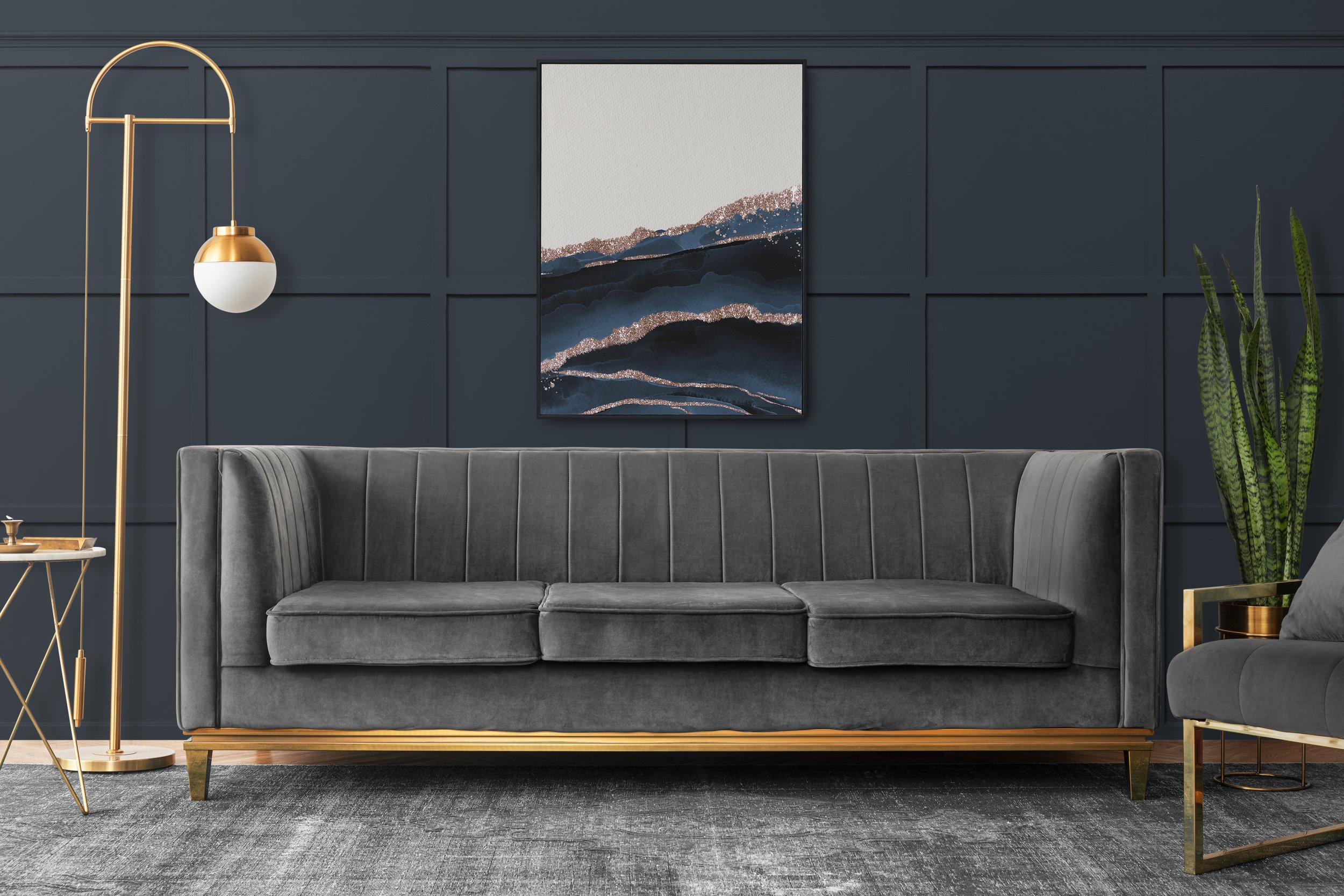 Modern living room with dark blue paneled wall, gray velvet sofa, abstract artwork, gold and white floor lamp, potted snake plant, and gray rug.