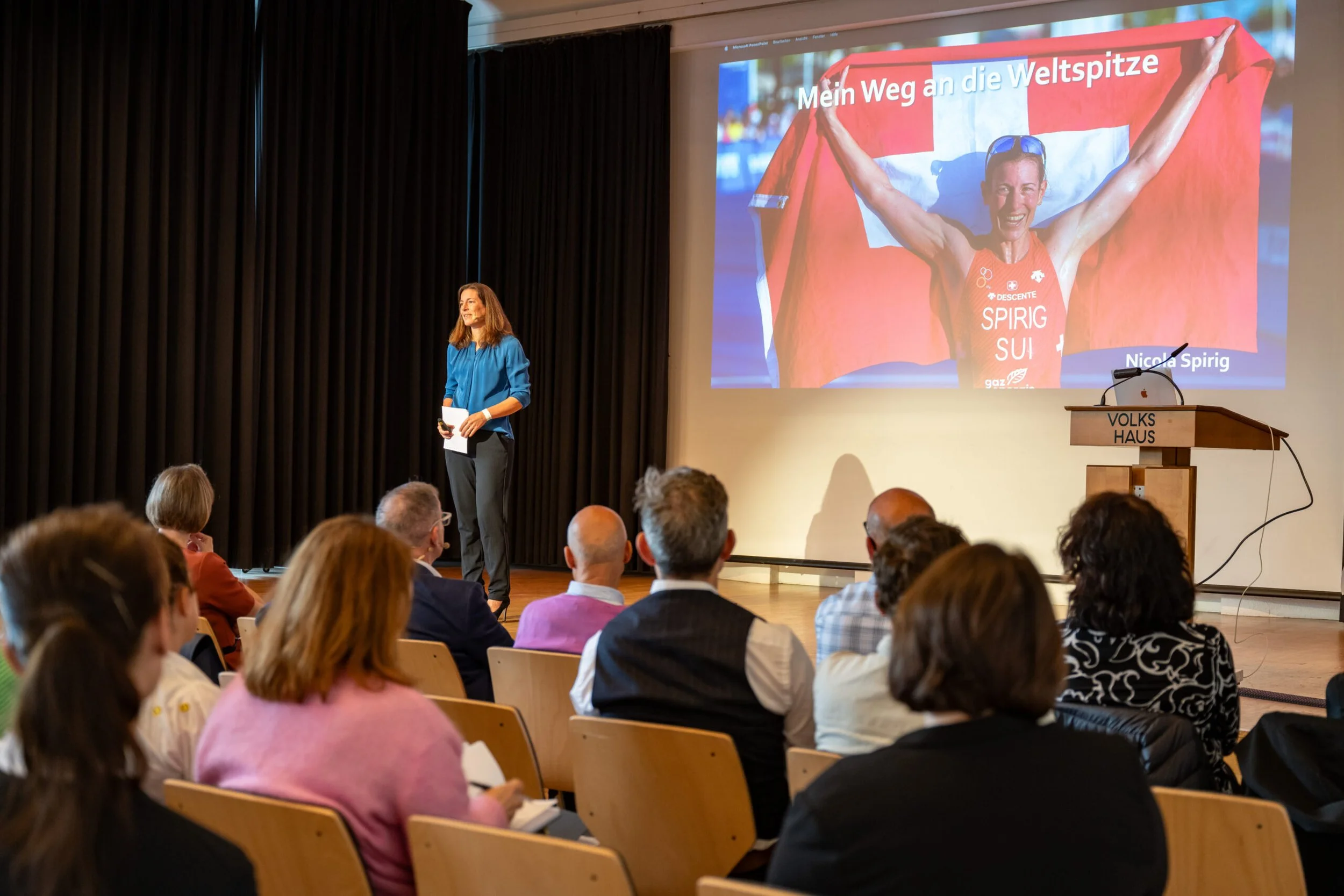 Eine Frau in blauer Bluse spricht vor einem Publikum in einem Saal. Auf der großen Leinwand ist ein Bild einer fröhlichen Athletin mit einer Schweizer Flagge im Hintergrund zu sehen, mit dem Text 'Mein Weg an die Weltspitze'.