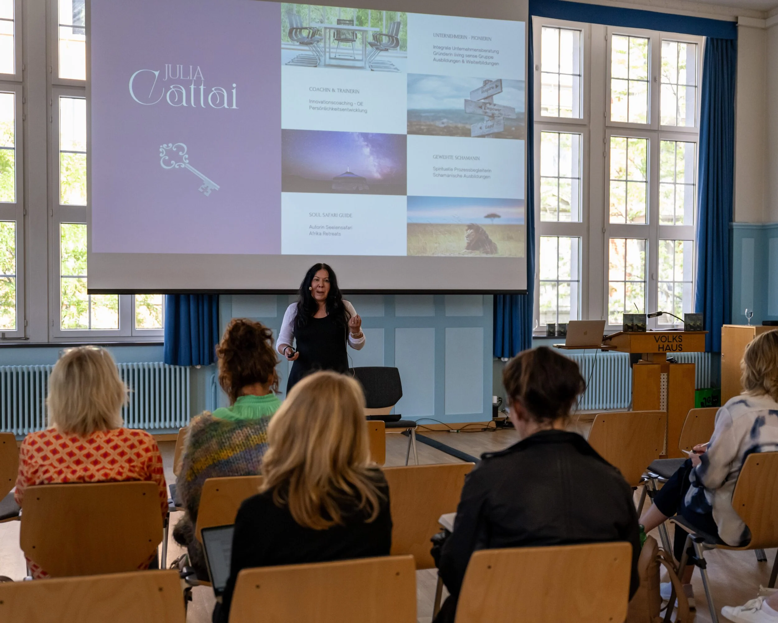 Eine Frau hält einen Vortrag vor einer Gruppe von Zuhörern in einem Raum mit großen Fenstern und blauer Wandverkleidung. Auf der großen Projektionswand ist eine Präsentation mit dem Titel 'JULIA VATTÁI', die verschiedene Bilder und Textfelder zeigt.