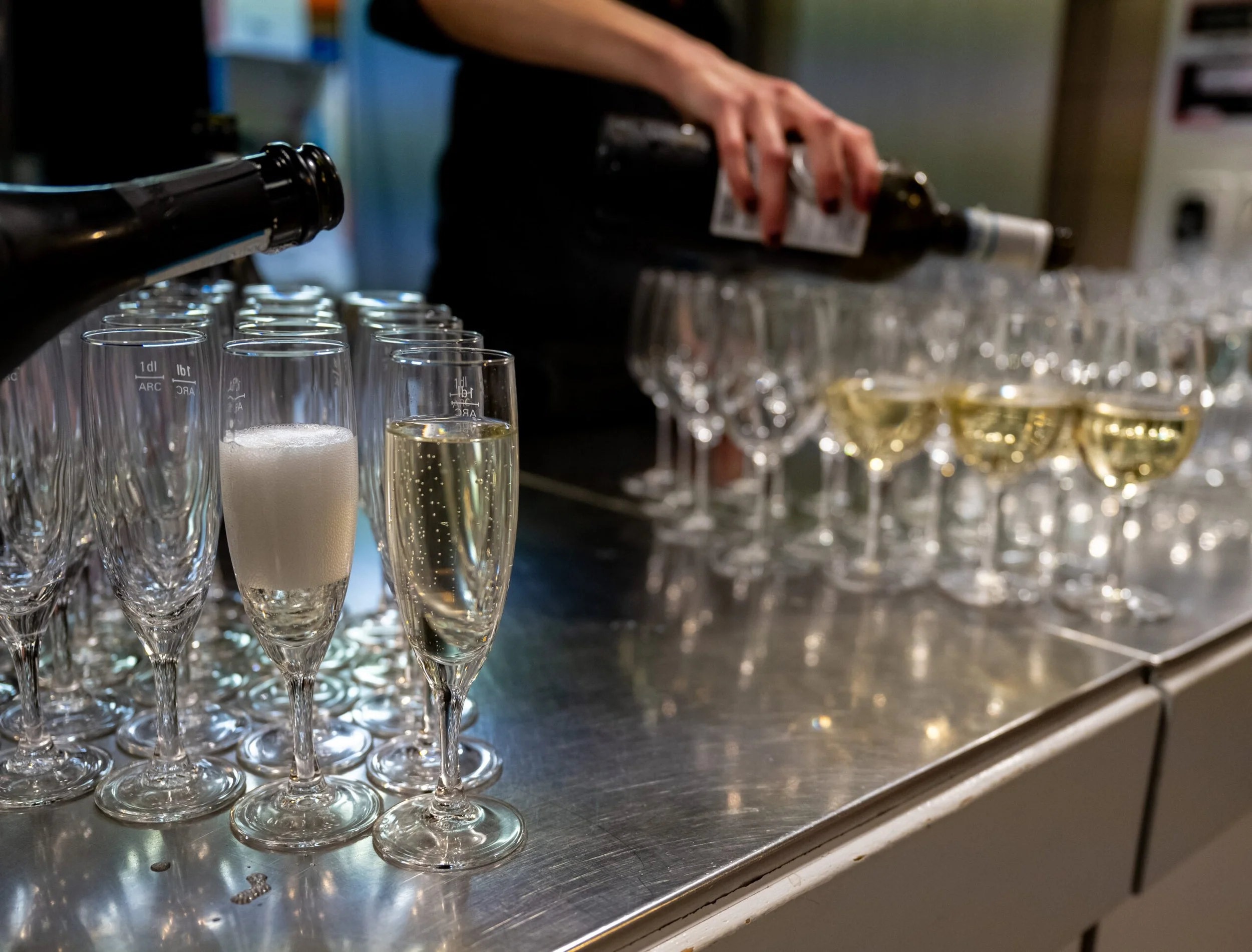 Mehrere Champagnergläser auf einem Tisch, einige sind gefüllt, andere sind leer. Jemand gießt einen Sekt oder Champagner in die Gläser.