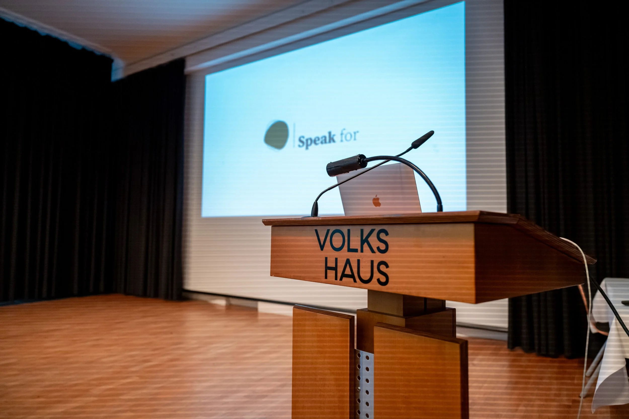 Foto eines Rednerpults mit der Aufschrift 'Volks Haus', einem Laptop und einem Mikrofon in einem Veranstaltungsraum, im Hintergrund eine große Leinwand mit einem Logo und dem Text 'Speak for'.