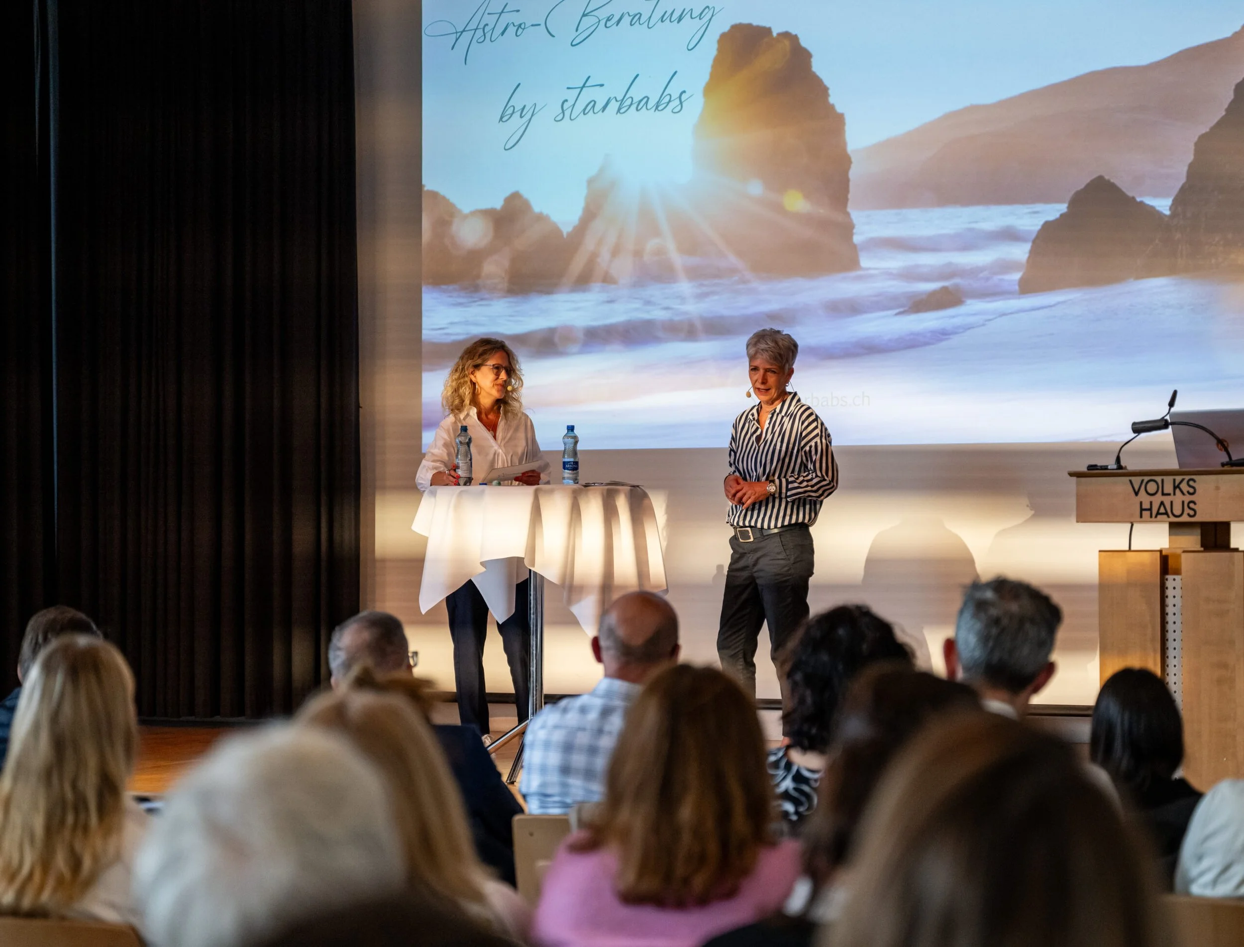 Zwei Frauen stehen an einem Mikrofon bei einer Präsentation vor Publikum, im Hintergrund ein großes Bildschirm mit Meereslandschaft und Sonnenaufgang, rechts ein Rednerpult mit der Beschriftung "Volks Haus".