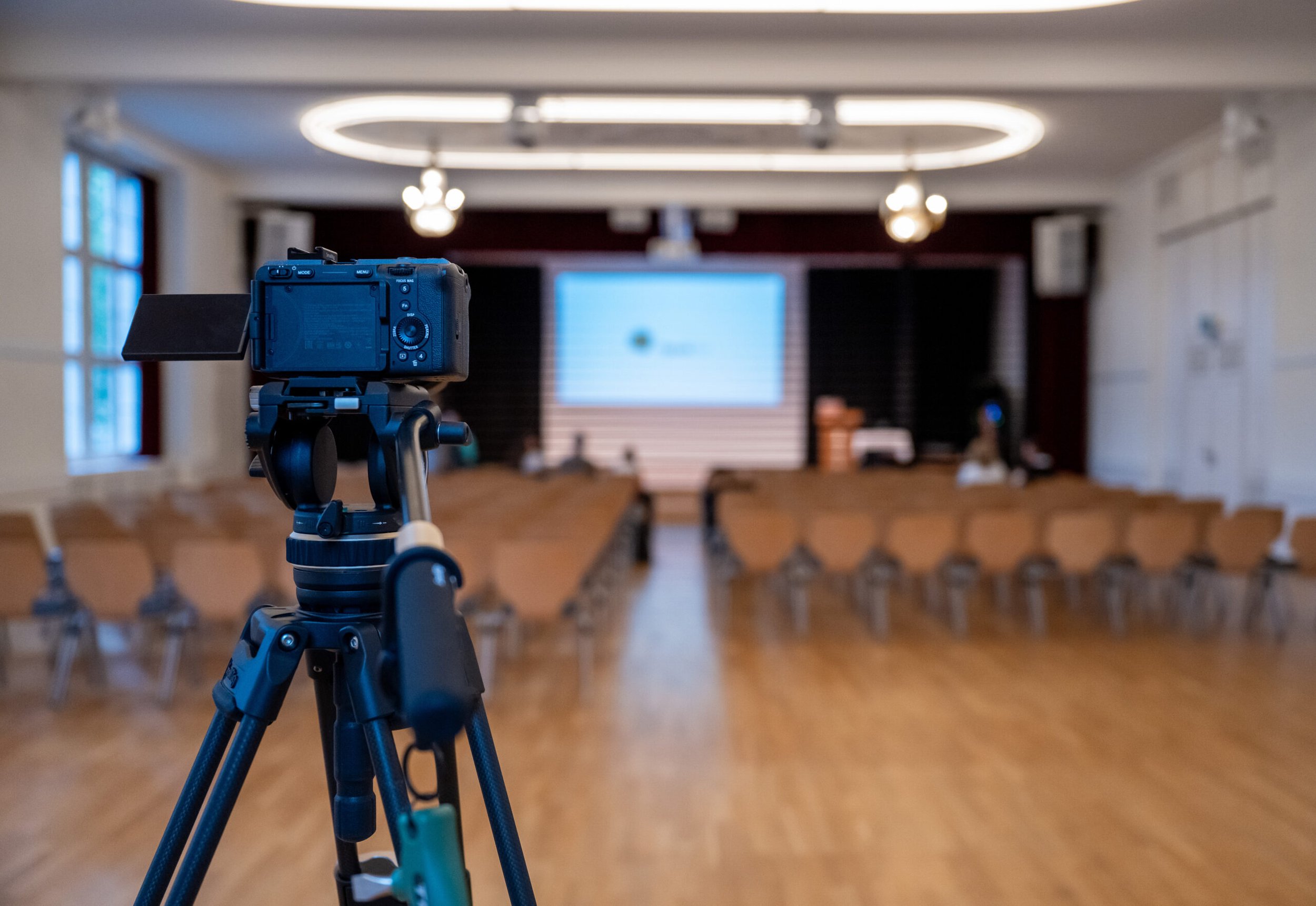 Aufnahmegerät auf Stativ in einem leeren Veranstaltungsraum mit Sitzreihen und einer großen Bildschirmanzeige an der Wand.