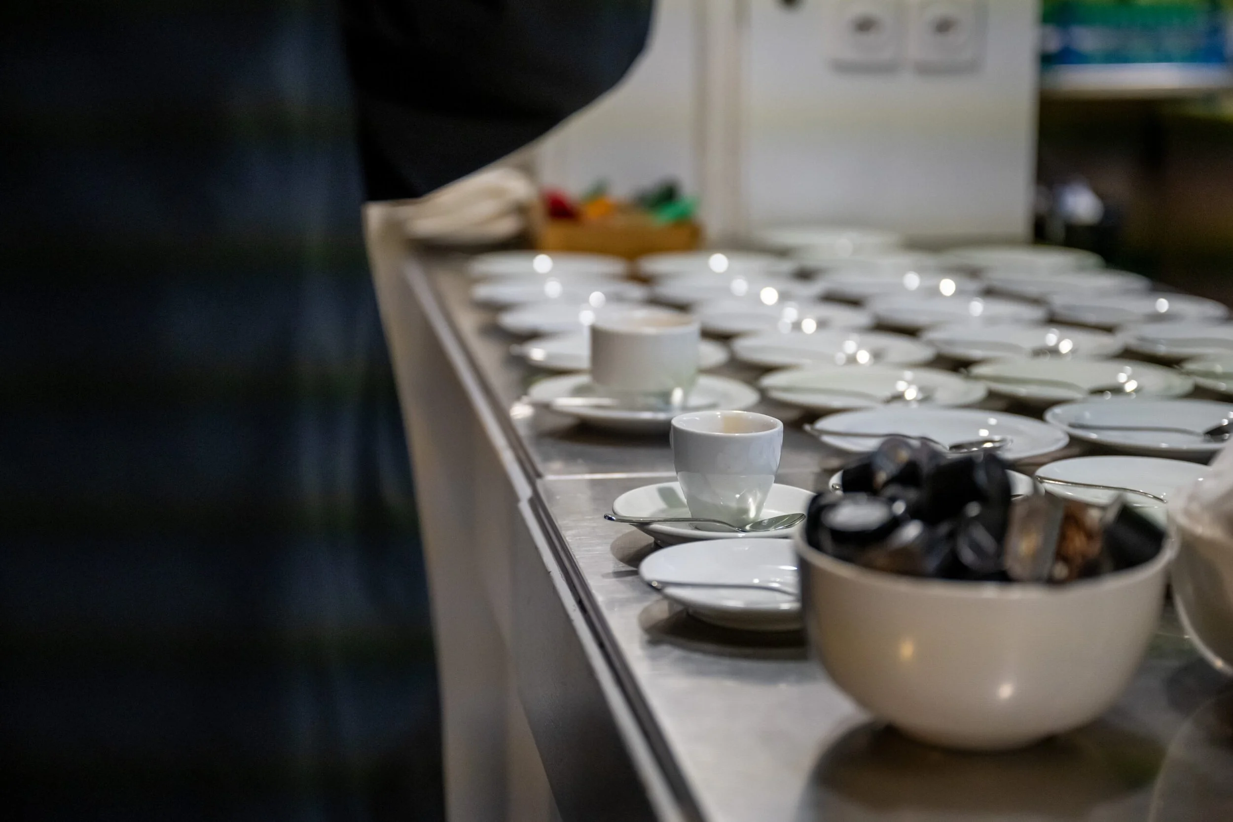 Leere weiße Kaffeetassen mit Untertassen und Löffeln auf einer Theke, in der Nähe eine große Schüssel mit schwarzen Kaffeekapseln.