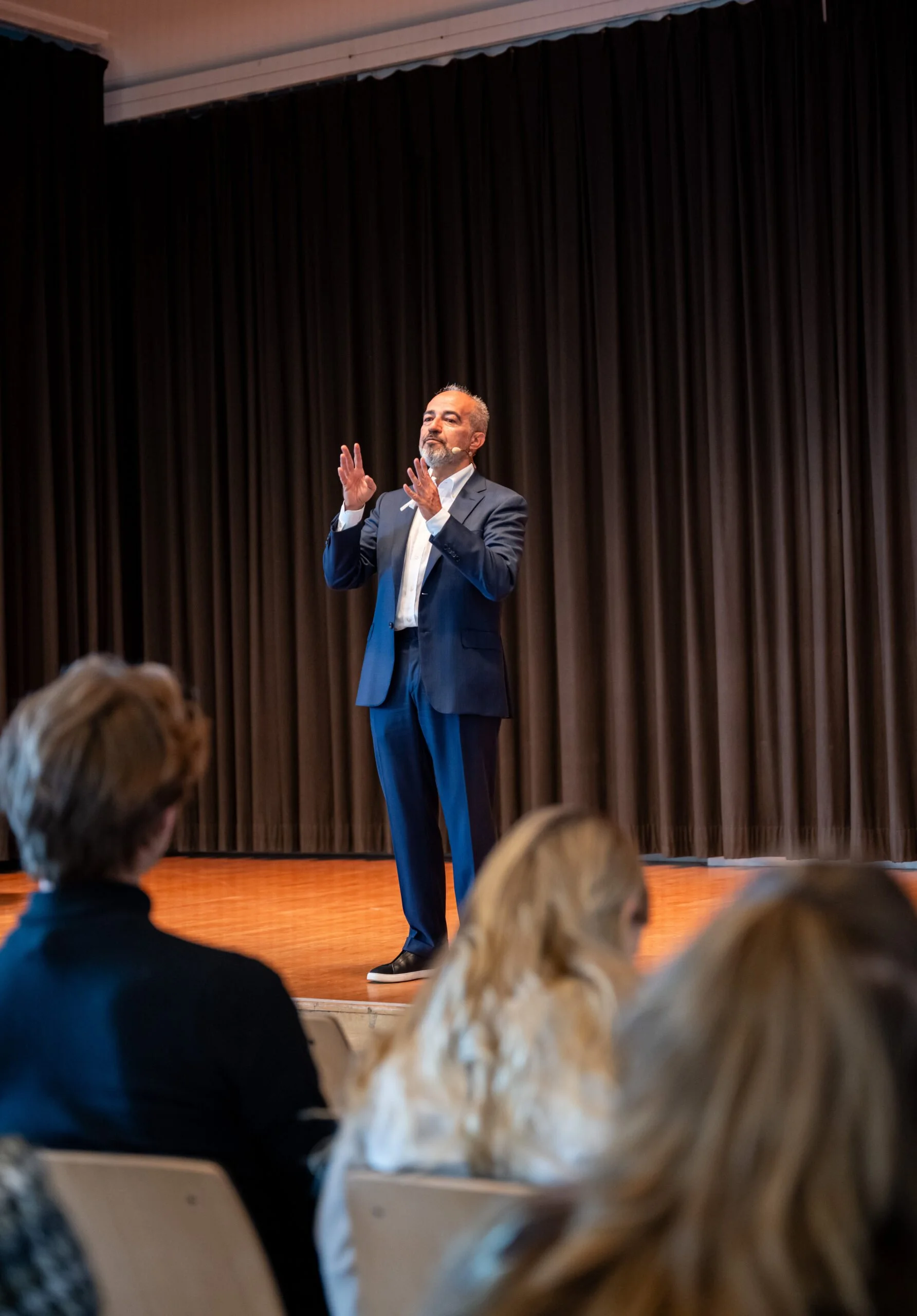 Mann in Anzug spricht auf Bühne vor einem dunklen Vorhang, Publikum hört aufmerksam zu.