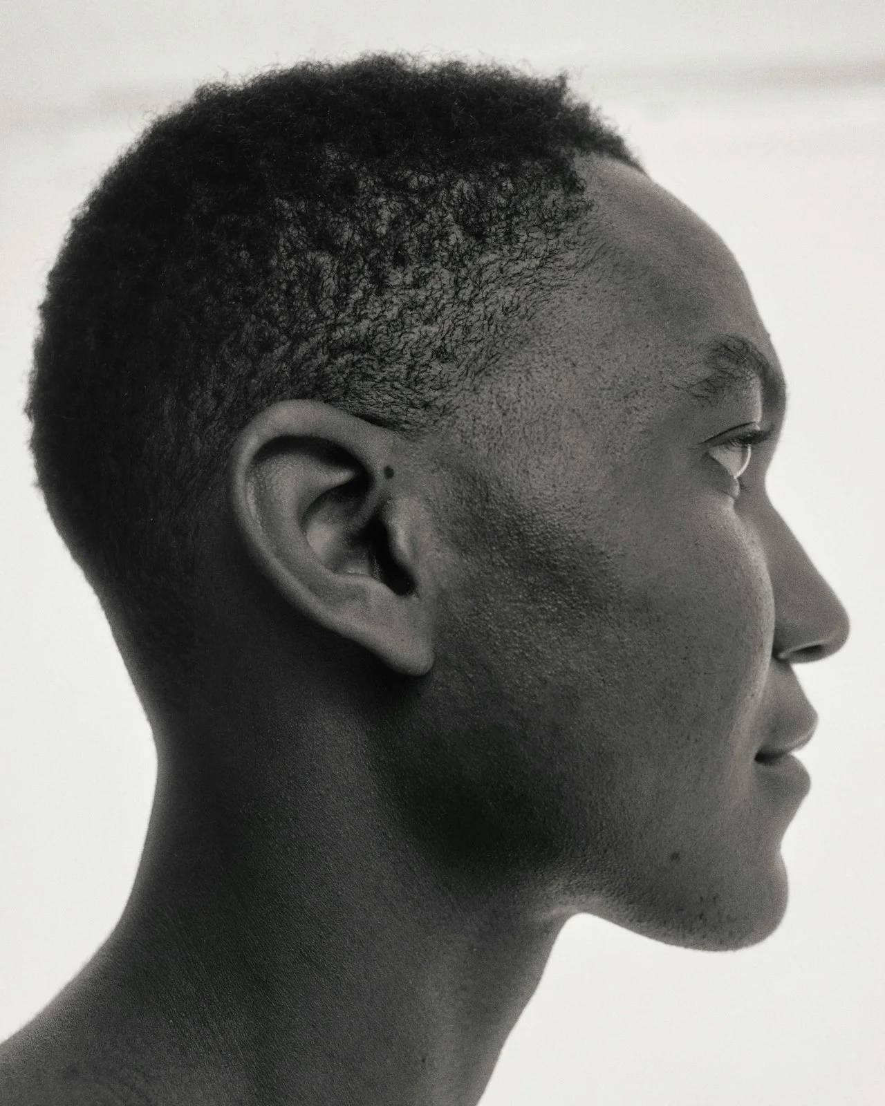 Side profile of a person's face with short curly hair and smooth skin, set against a plain background.