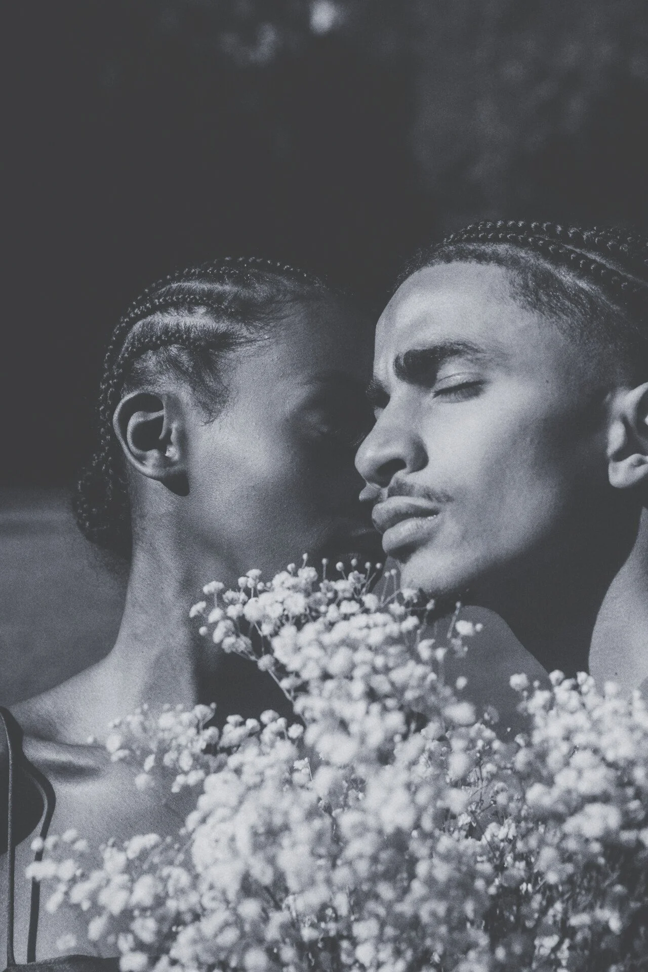 Black and white photo of a couple with braided hair, holding a bouquet of small flowers, close to each other with eyes closed.