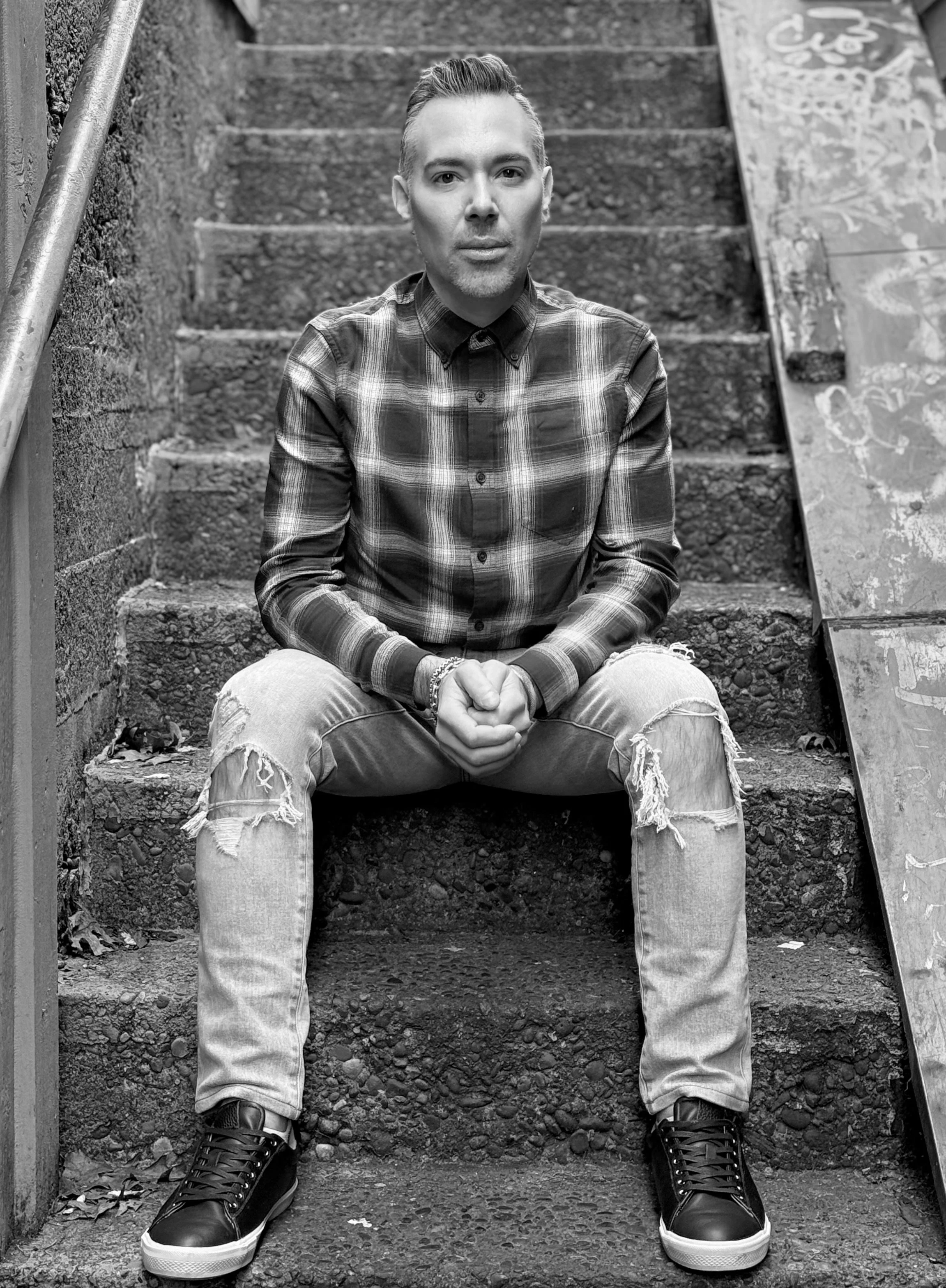 A man sitting on outdoor concrete stairs, wearing a plaid shirt, ripped jeans, and sneakers, with a hand bracelet, looking directly at the camera. The setting appears urban with graffiti on a nearby sign.