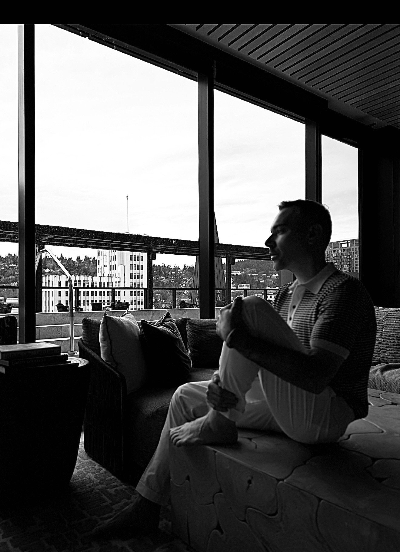 Black and white photo of a man sitting on a bed or sofa near large windows, holding a blanket or pillow, with an urban cityscape visible outside.