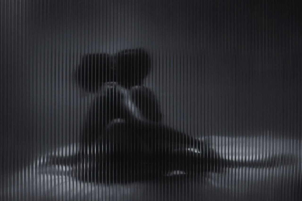 Silhouette of a person sitting on the ground behind a semi-transparent ribbed glass surface.