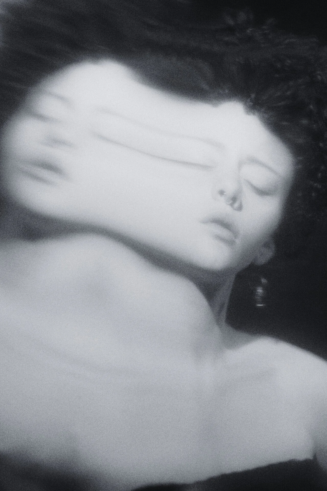 A black-and-white photograph of a woman with long hair, with her eyes closed and her hand covering half of her face.