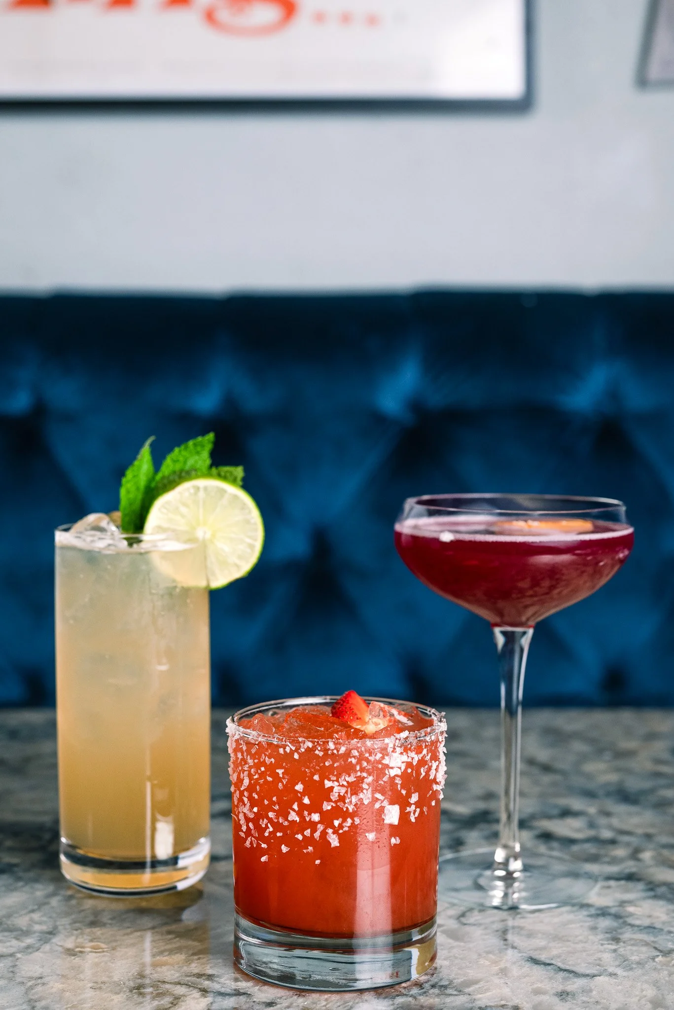 Three colorful cocktails on a marble table against a dark blue, tufted banquette. The left drink is a pale yellow, garnished with a lime slice and mint leaves. The middle drink is an orange-red, rimmed with salt and topped with a strawberry. The righ