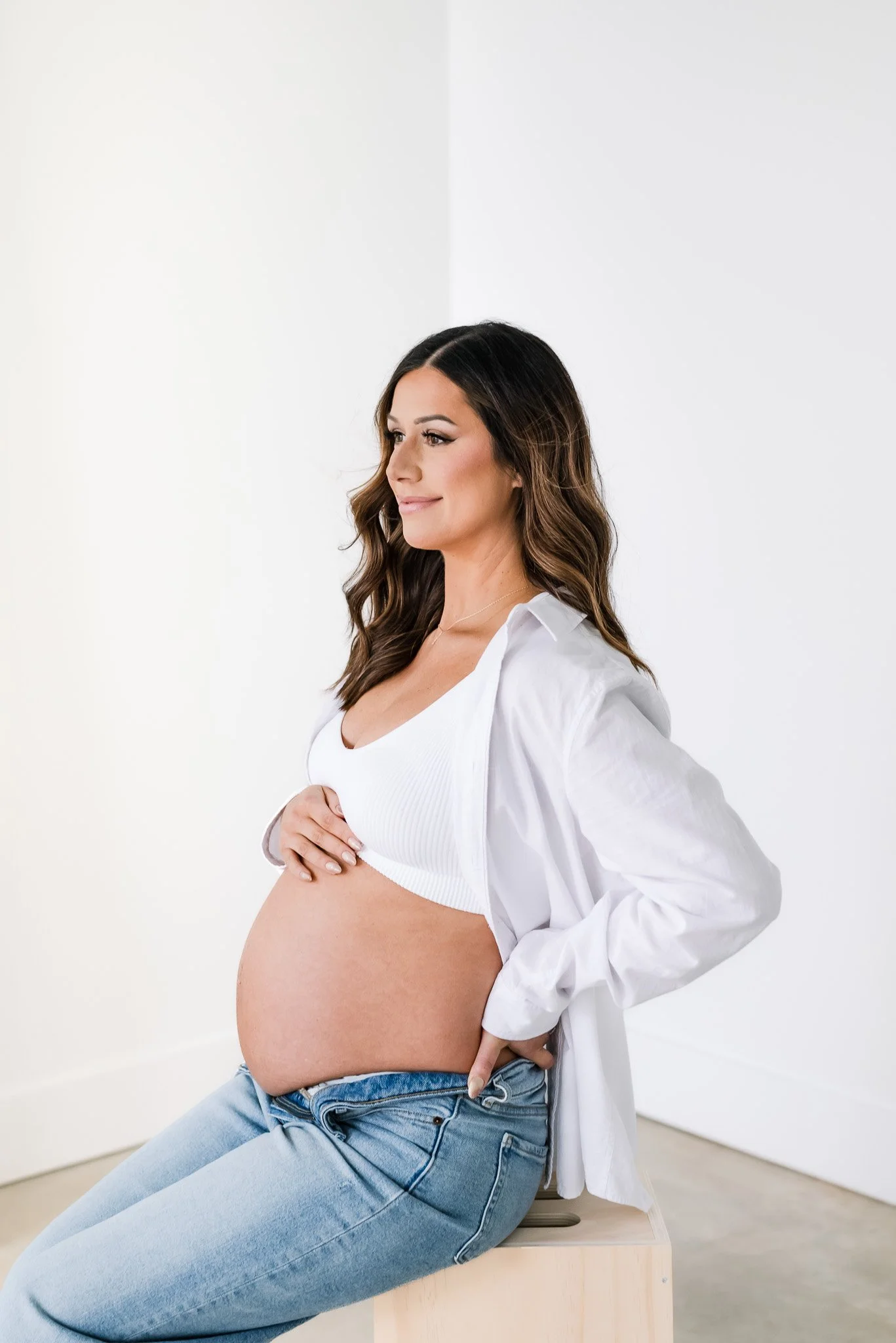 Pregnant woman sitting on a wooden stool, wearing a white tank top and unbuttoned white shirt, with her hand on her belly and looking to the side, in a bright room.