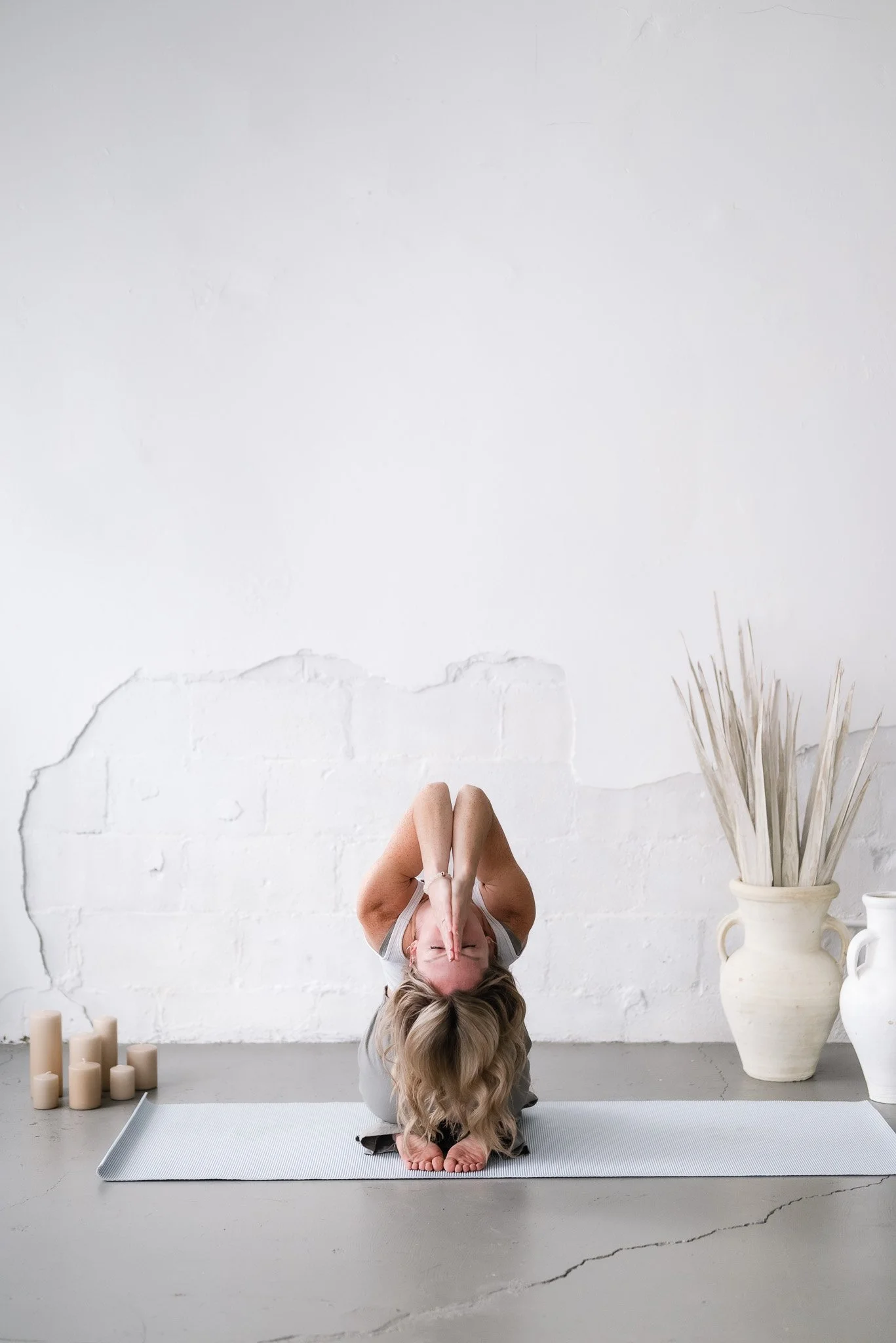A woman practicing yoga in a kneeling forward bend pose on a mat in a minimalist room with candles and large vases.