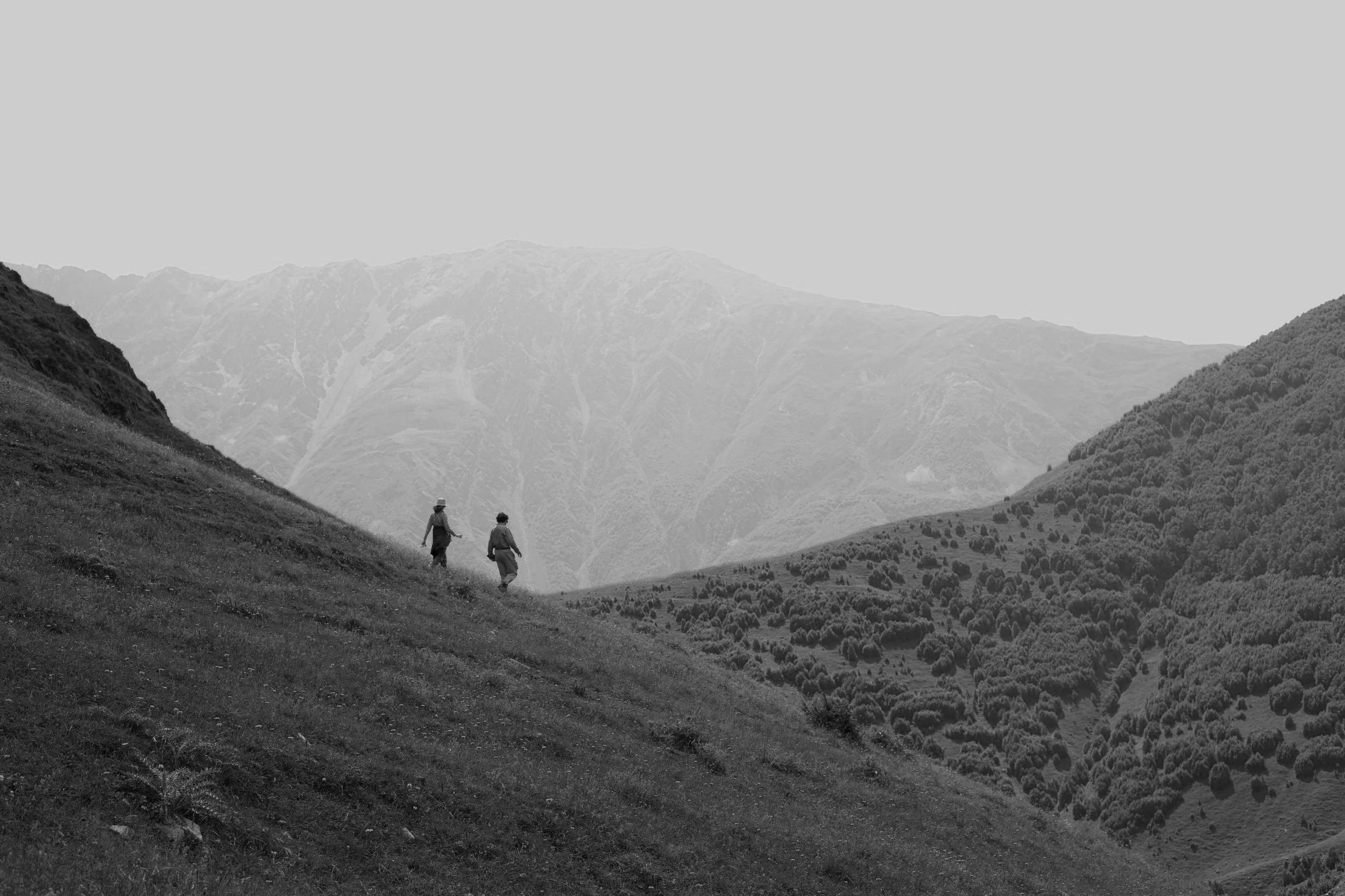 Two people hiking in a mountainous landscape with rolling hills and mountain peaks in the background.