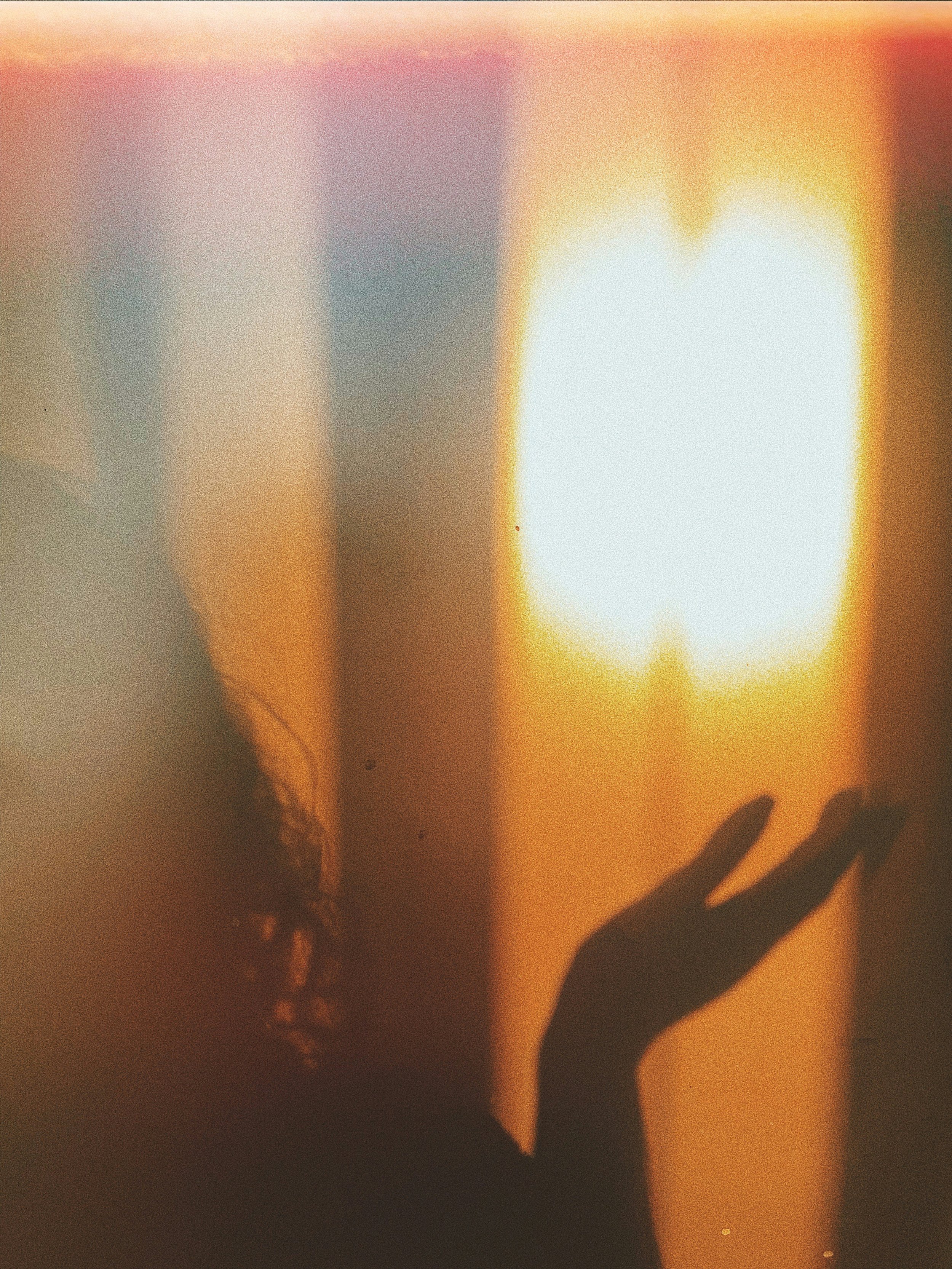 A hand silhouette reaching out towards a blurred, bright light source in a dark room, with warm orange and yellow hues.