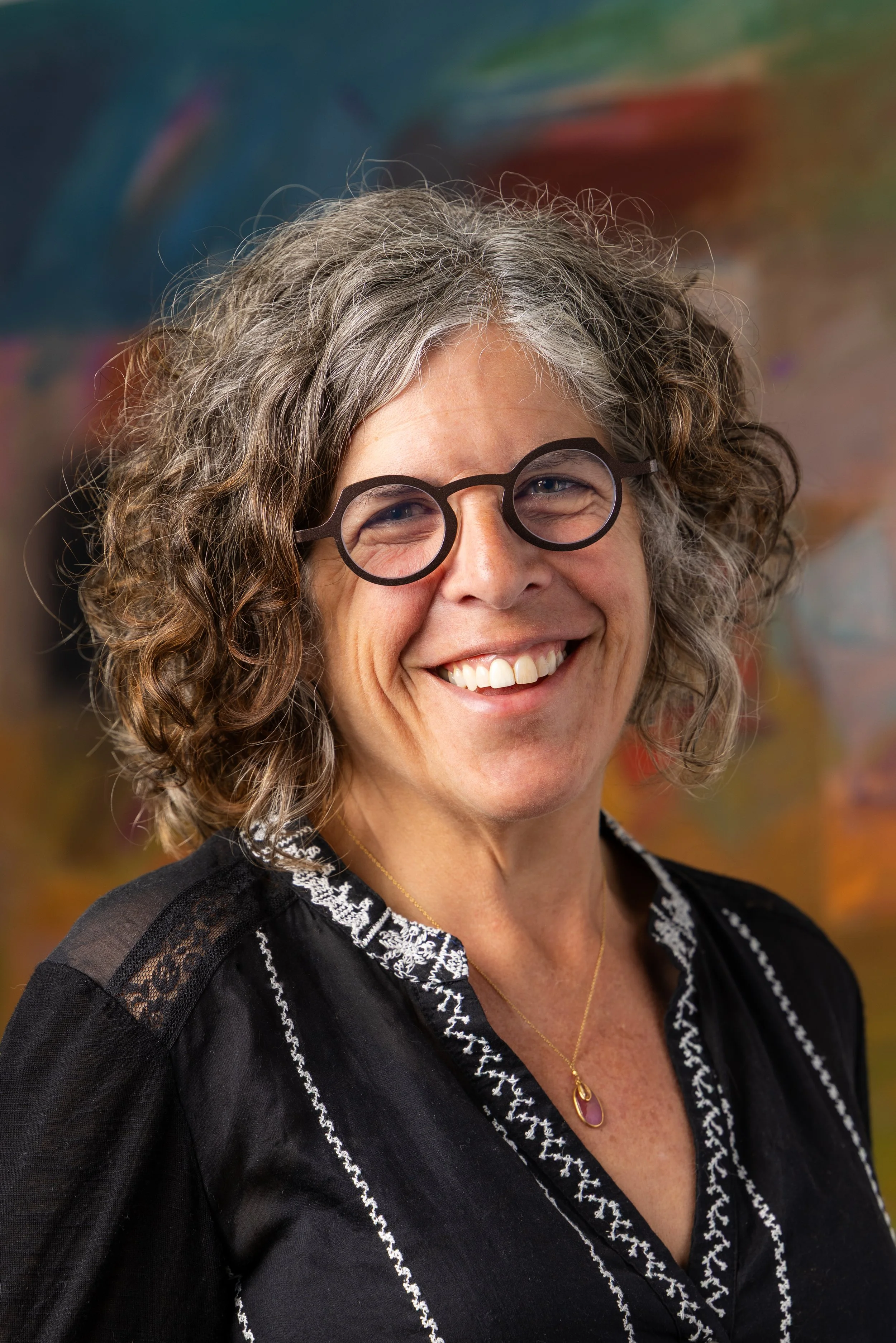 Close-up of a smiling woman with grey, curly hair wearing glasses, a black top with lace and embroidery, and a gold necklace, against a colorful, blurred background.