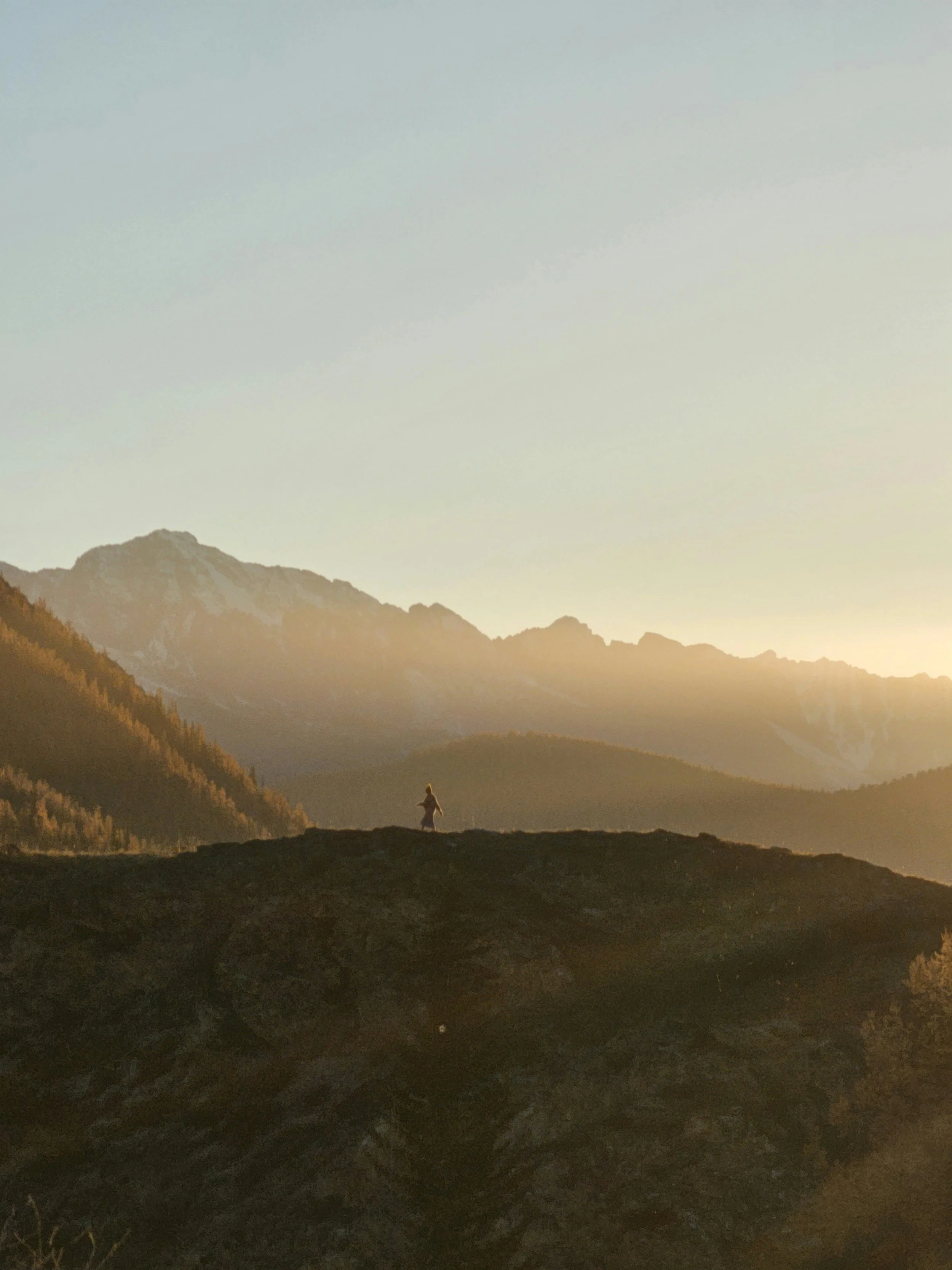 A person standing on a hilltop with mountains in the background at sunset.