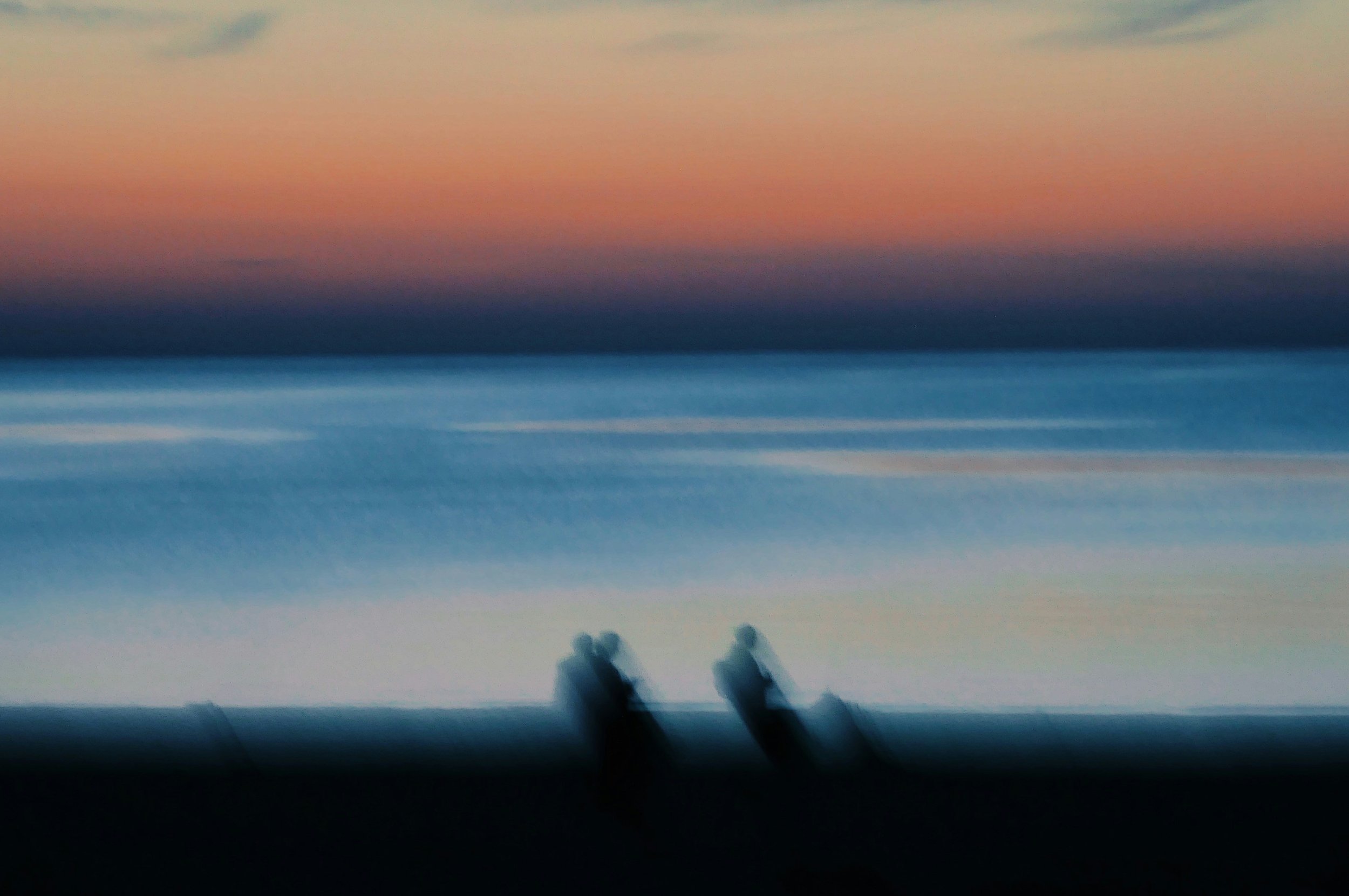 Blurred image of a sunset over the ocean with silhouettes of two people standing on the beach in the foreground.