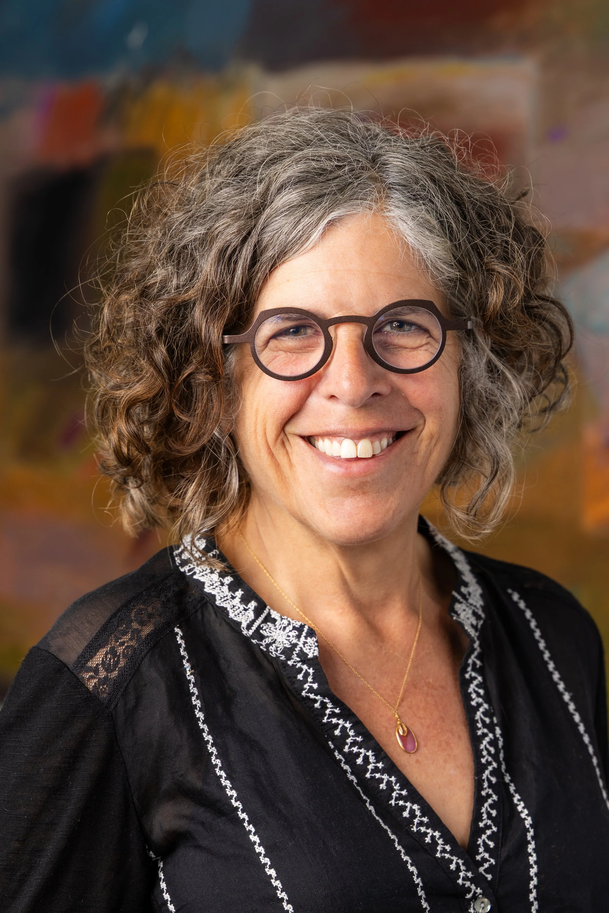A smiling woman with gray curly hair, wearing round glasses, a black embroidered top, and a gold necklace with a pink pendant. The background is blurred with various colors.
