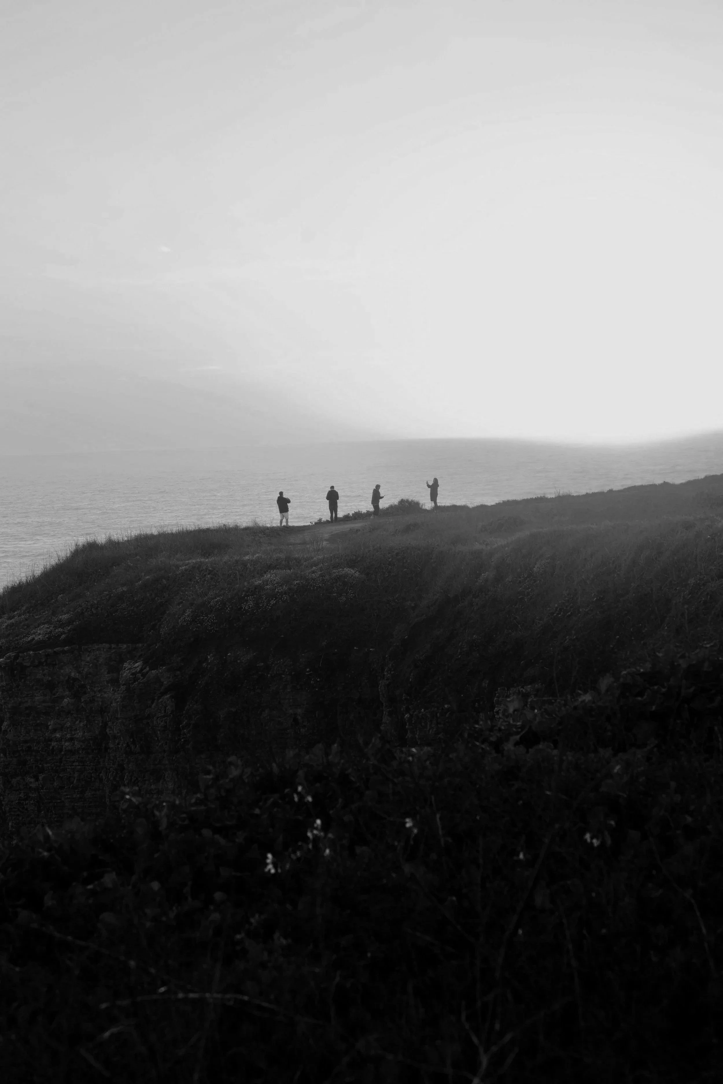 Black and white photo of four people standing on a grassy hill near the ocean, with the sky and water in the background.