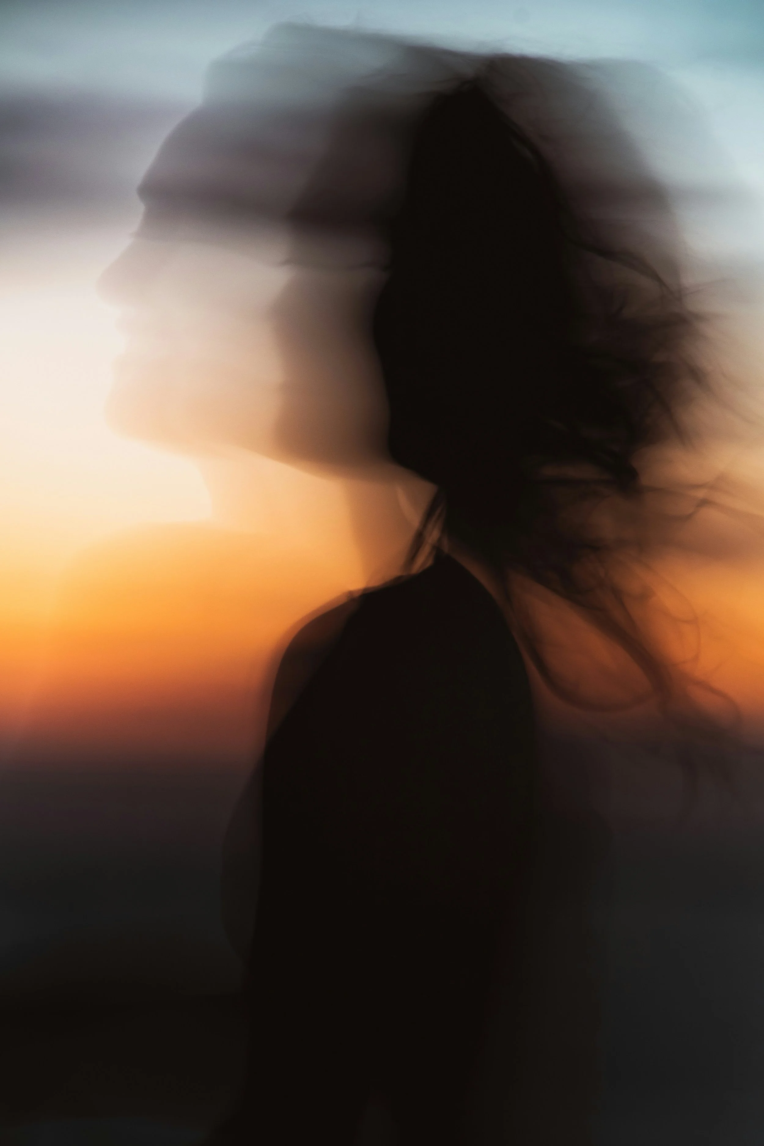 A silhouette of a woman with flowing hair standing against a sunset or sunrise sky, creating a peaceful and contemplative mood.