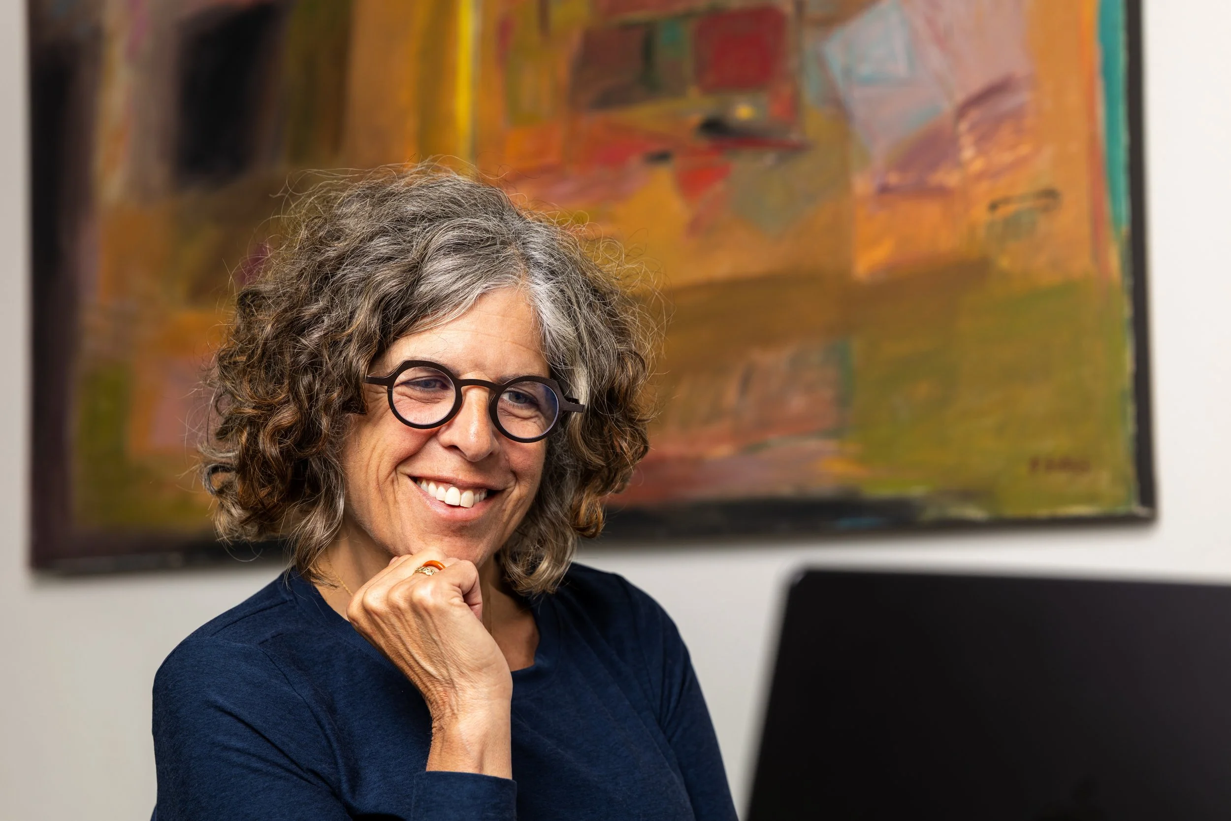 A woman with curly gray hair and glasses smiling, standing in front of an abstract painting in a gallery or museum.