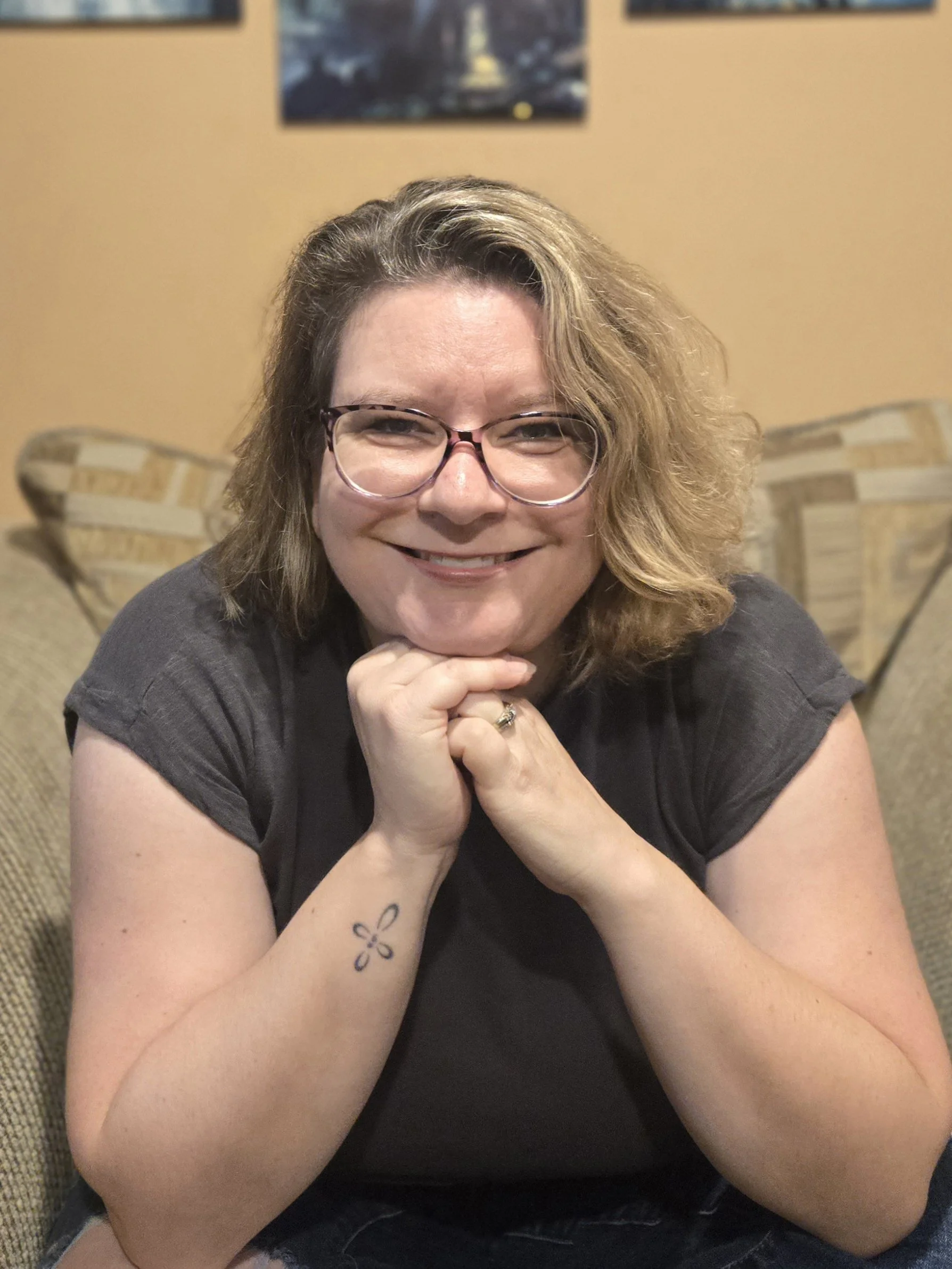 A woman with glasses and wavy blonde hair smiling while sitting on a couch.