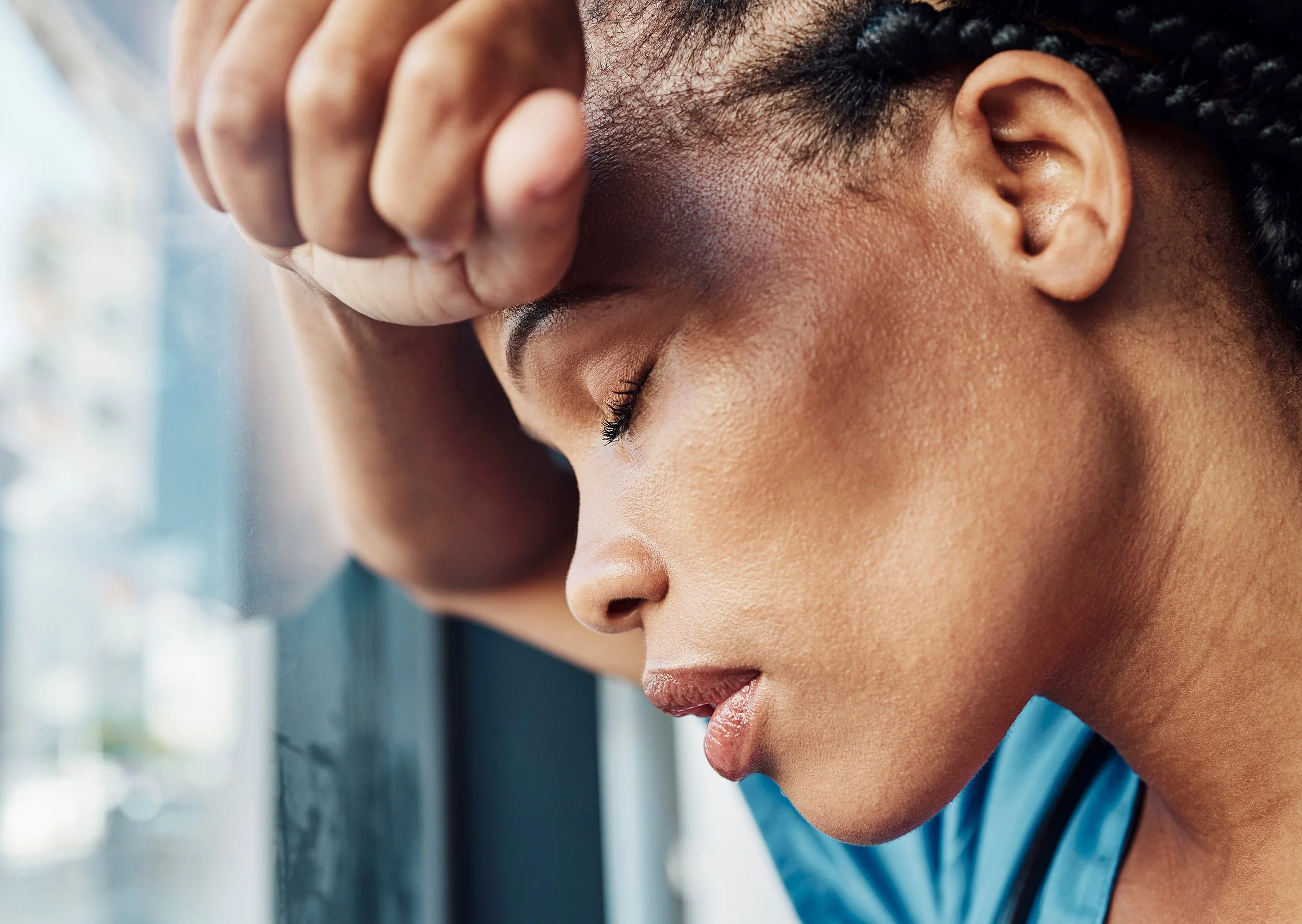close up of a sad woman with hand on forehead leaning against a window