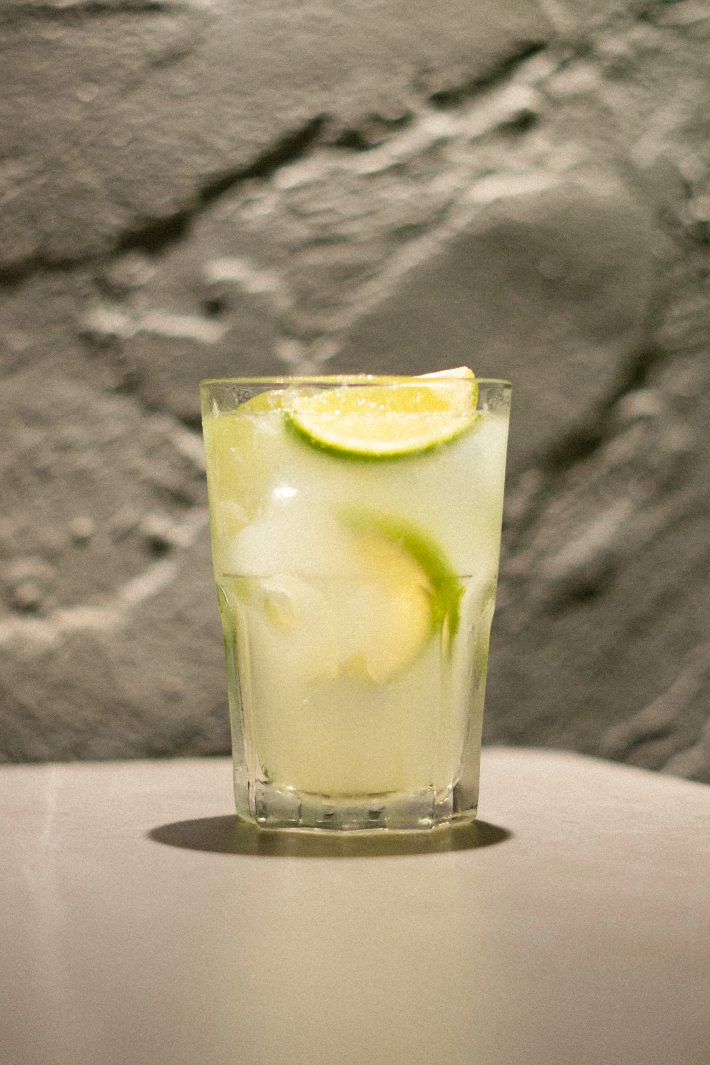 A glass of lemonade garnished with lime wedges on a white surface with a textured brick wall background.
