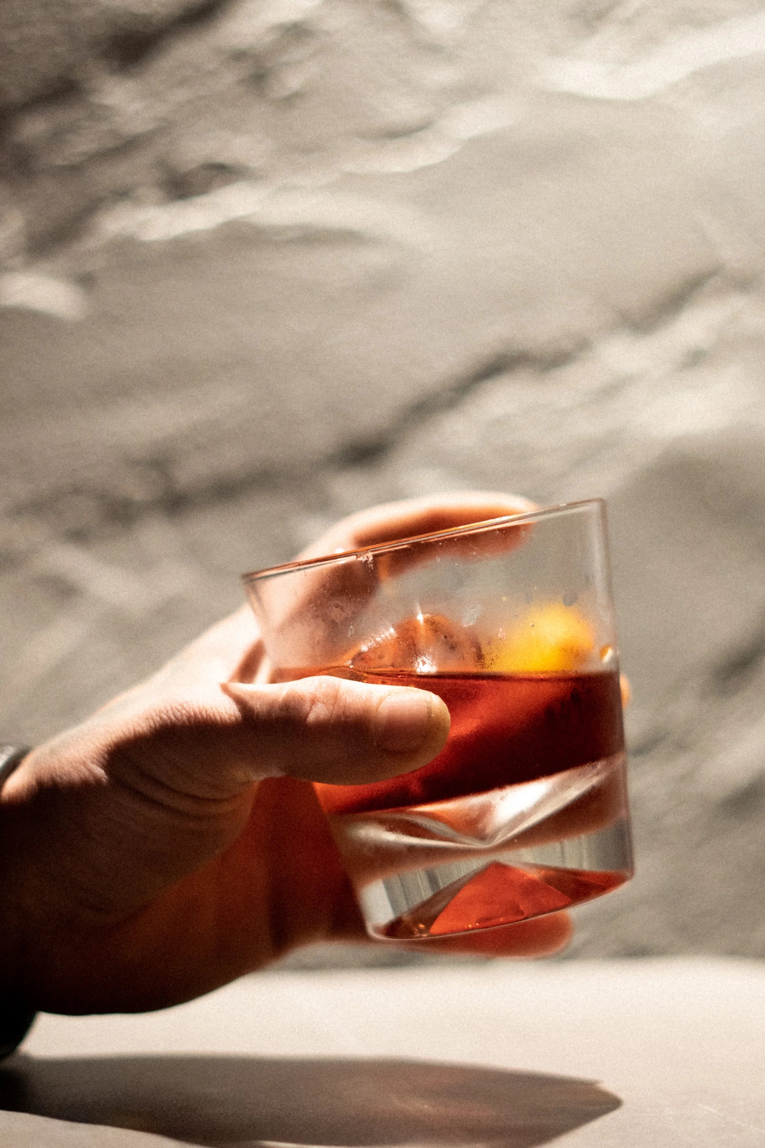 A hand holding a glass of red-colored beverage with ice, against a blurred outdoor background.