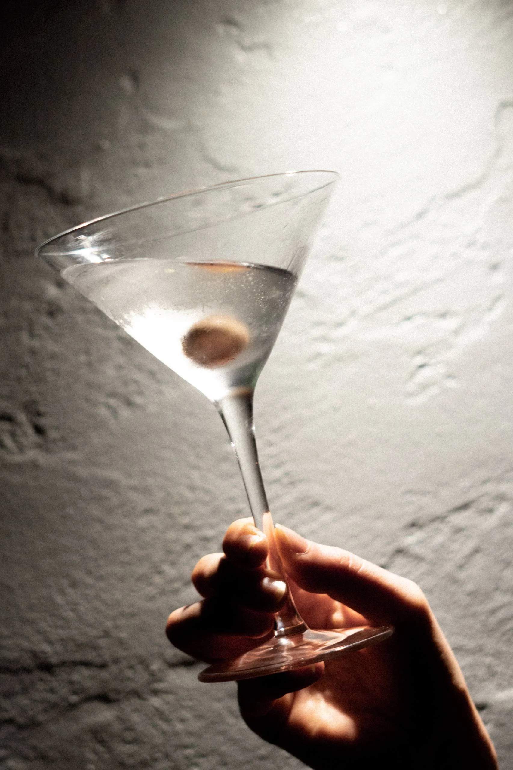 Hand holding a martini glass with an olive inside, against a textured light-colored wall.