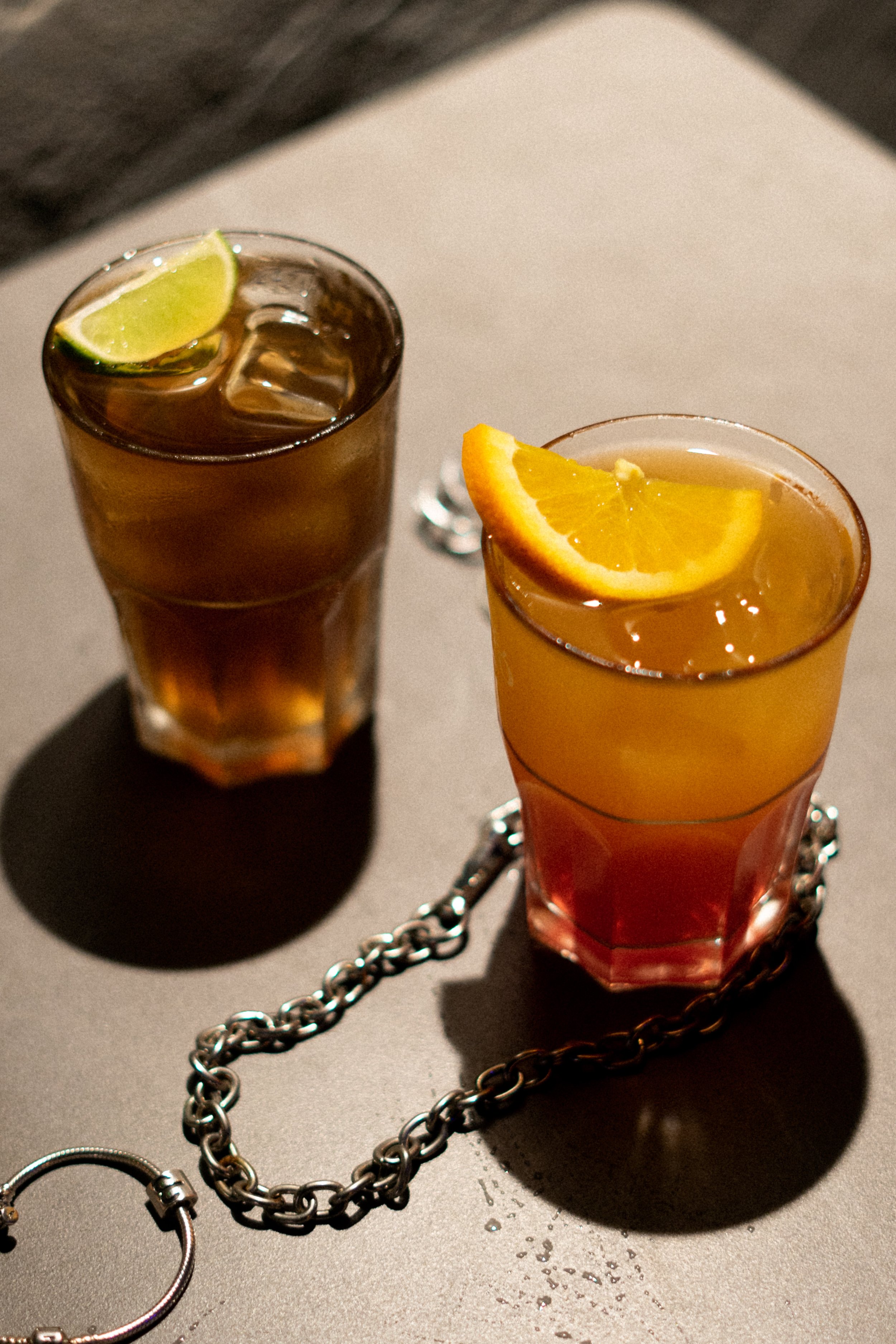 Two glasses of iced tea with lemon and lime wedges, connected by a chain, on a gray surface.