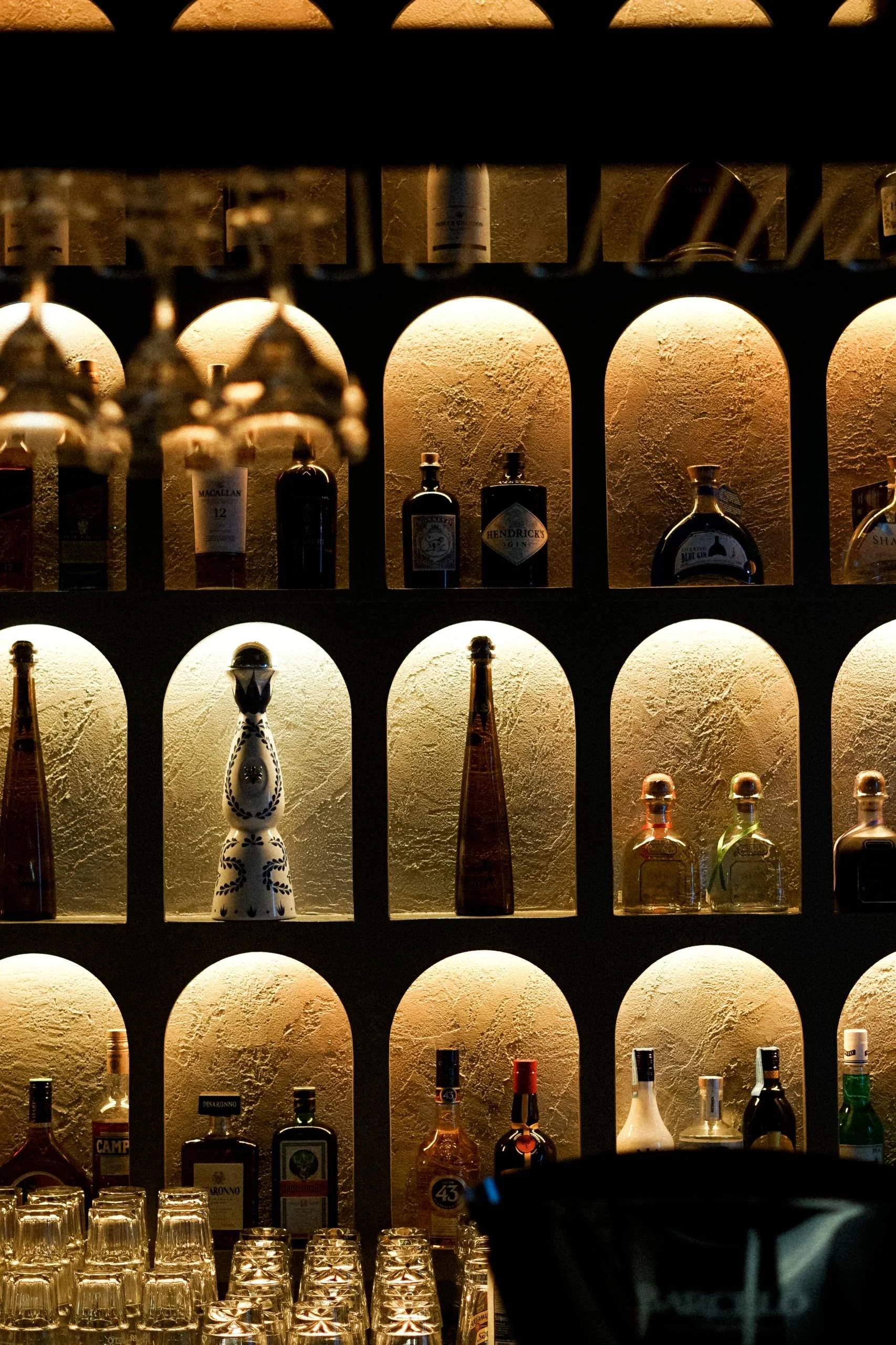 Shelf with liquor bottles illuminated by warm lighting, glasses at the bottom, and a black object partially visible in the foreground.