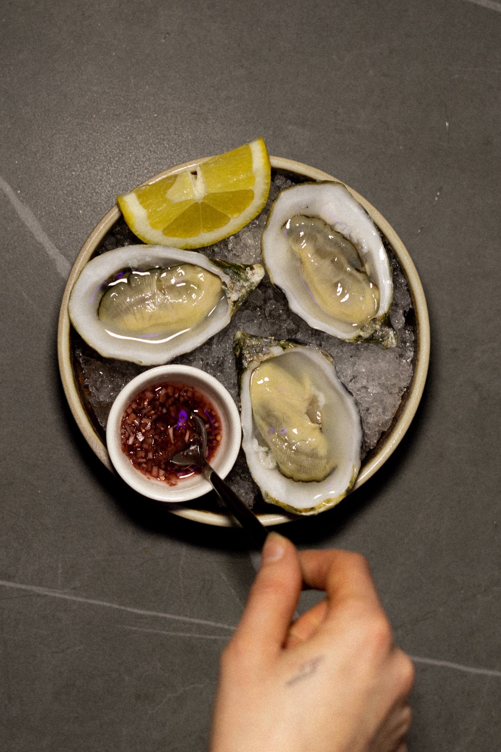 A round plate of oysters on ice with lemon wedge and dipping sauce.