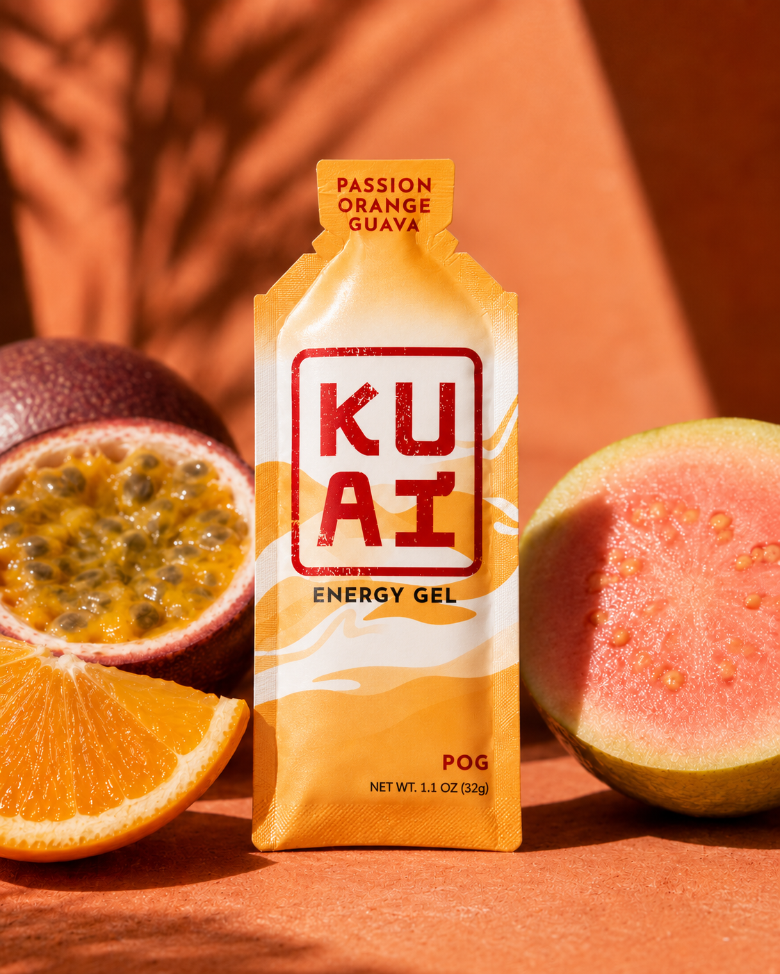 A KUAT energy gel packet with passion orange guava flavor, placed among half a passion fruit, a sliced orange, and a half-cut guava on an orange surface.