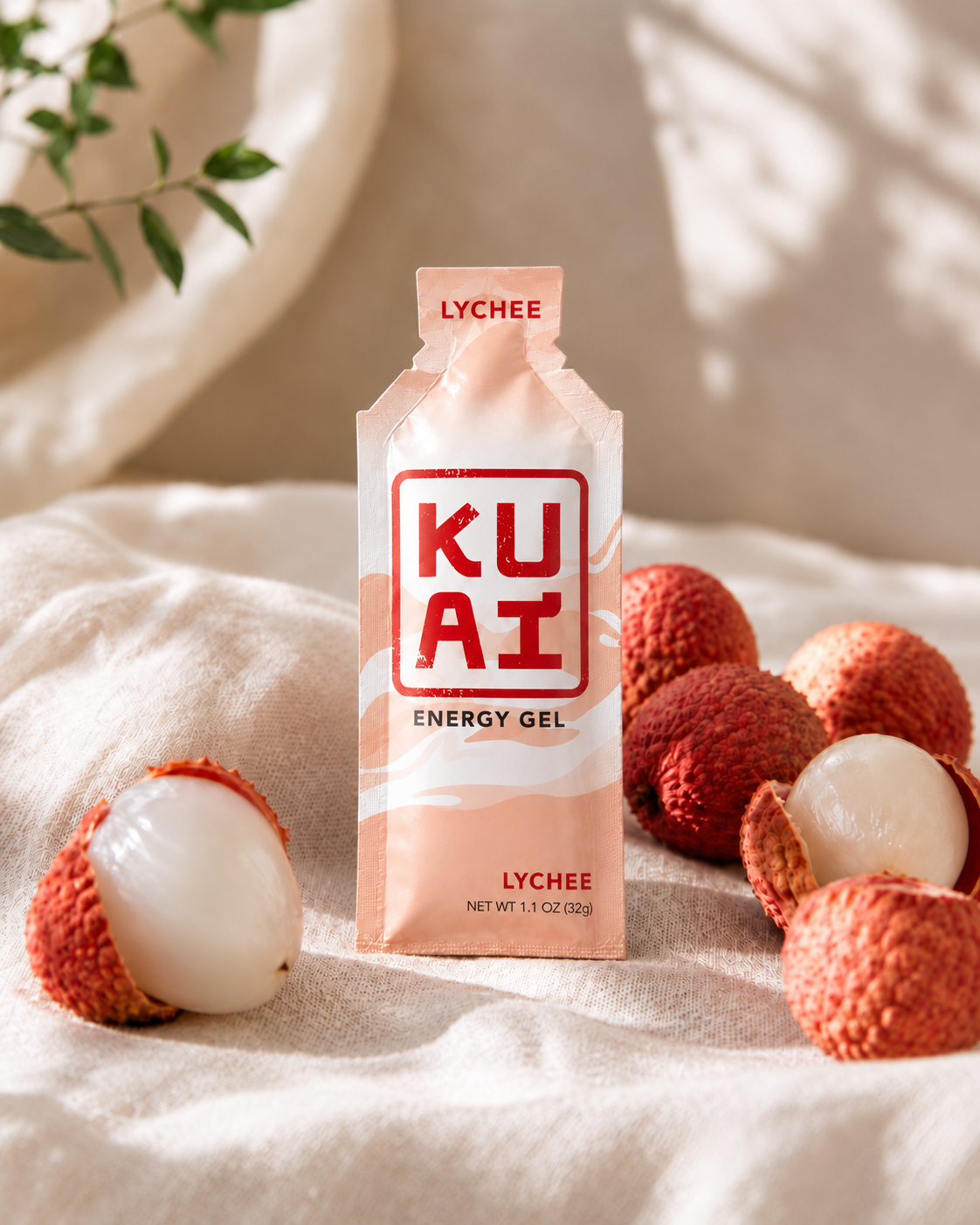 A lychee flavored energy gel packet labeled 'KUA'I' standing upright on a soft surface. Surrounding the packet are fresh lychee fruits, some peeled to reveal the white flesh inside.