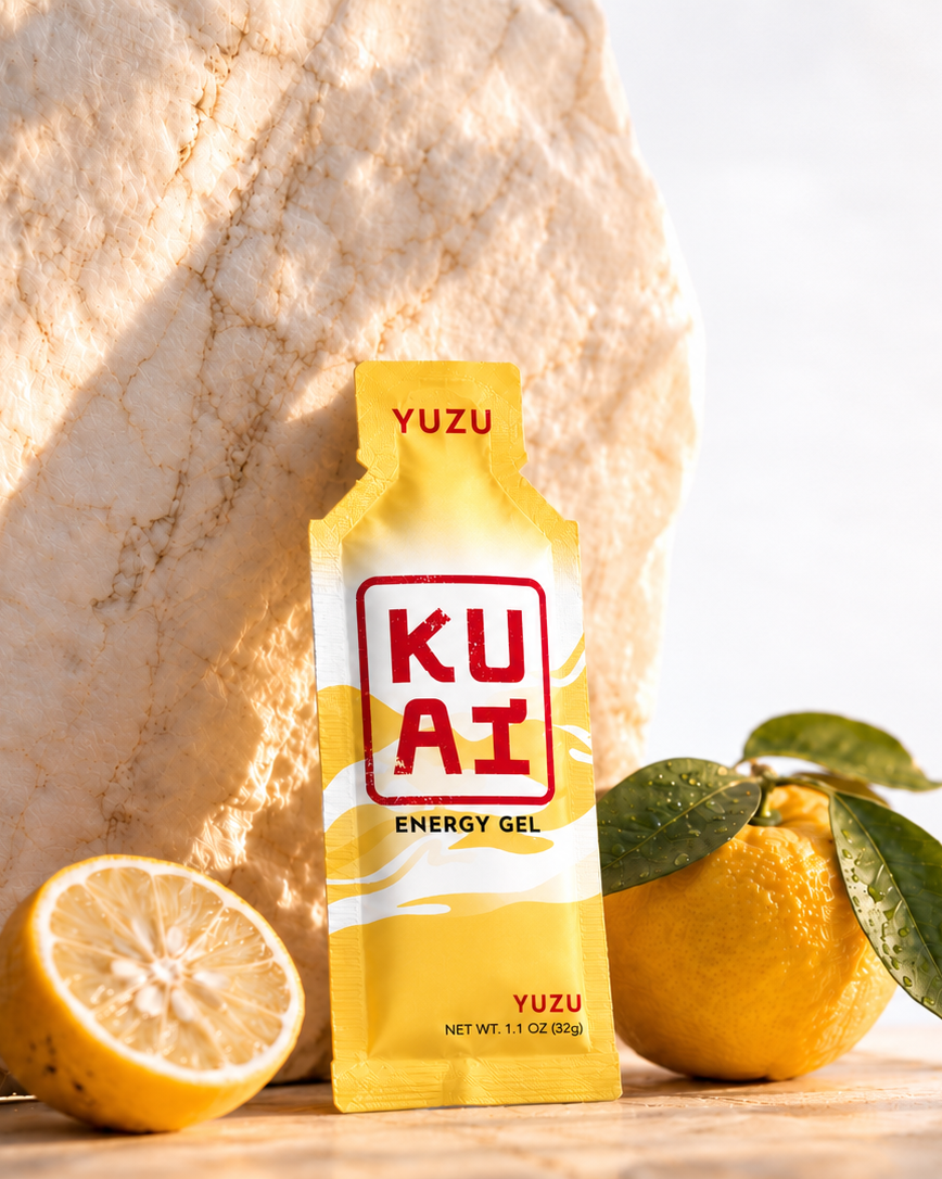 A yellow packet of KUAI energy gel with yuzu flavor, placed in front of a lemon and a large textured mineral stone, with lemon leaves and a whole lemon in the background.