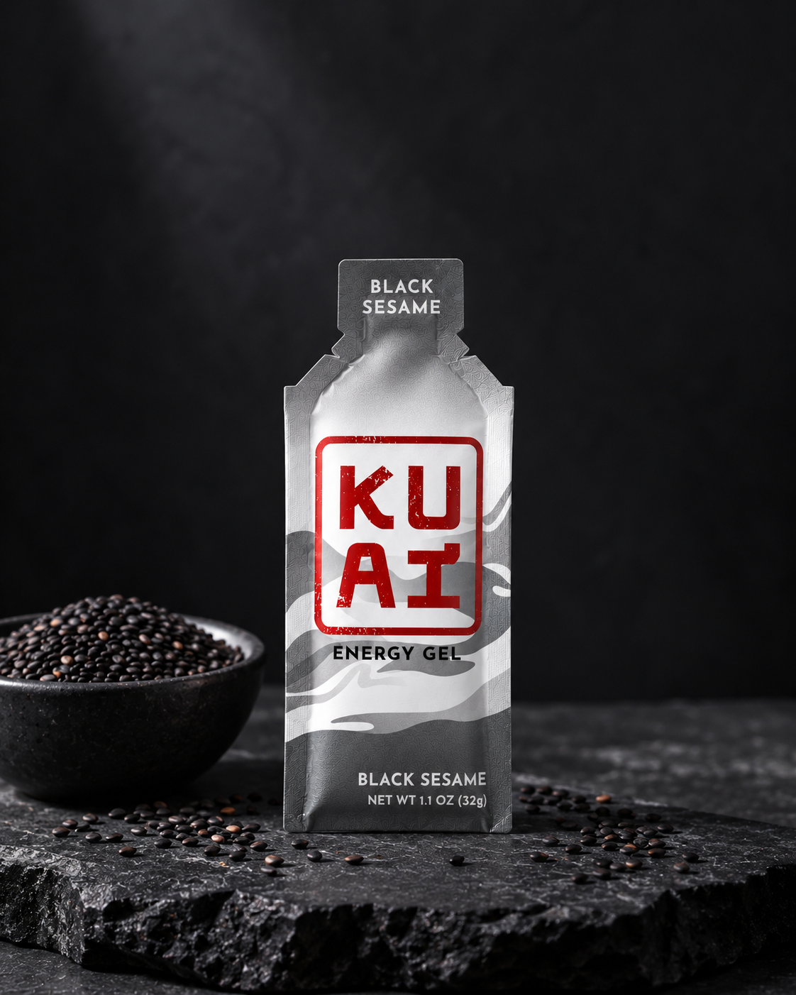 A packet of KUAT energy gel with black sesame flavor standing on a dark textured surface, next to a black bowl filled with black sesame seeds, all against a black background.