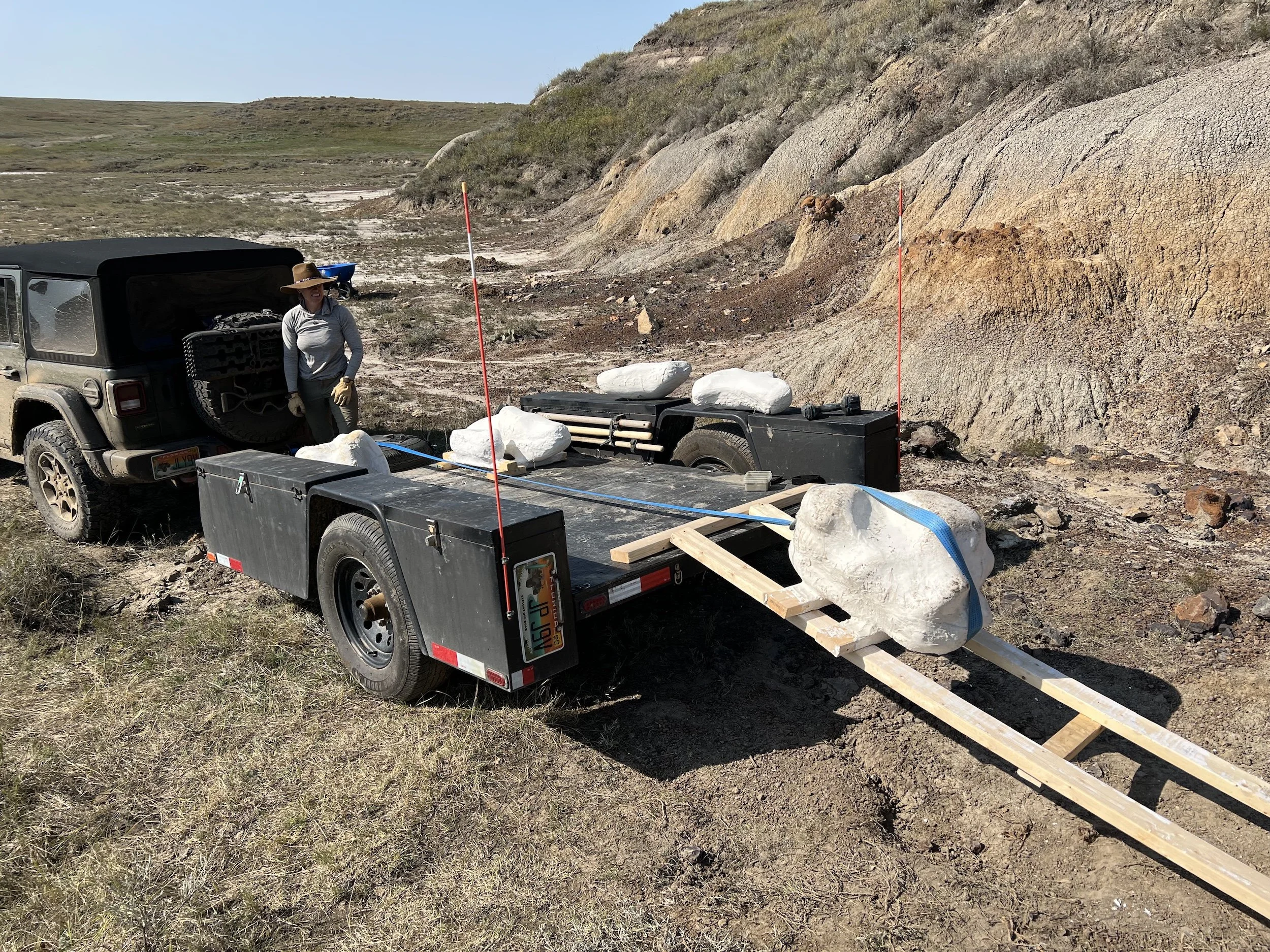 A person standing next to a black utility trailer with large rocks loaded on it, attached to a black vehicle, in a rugged outdoor landscape with dirt and hills.