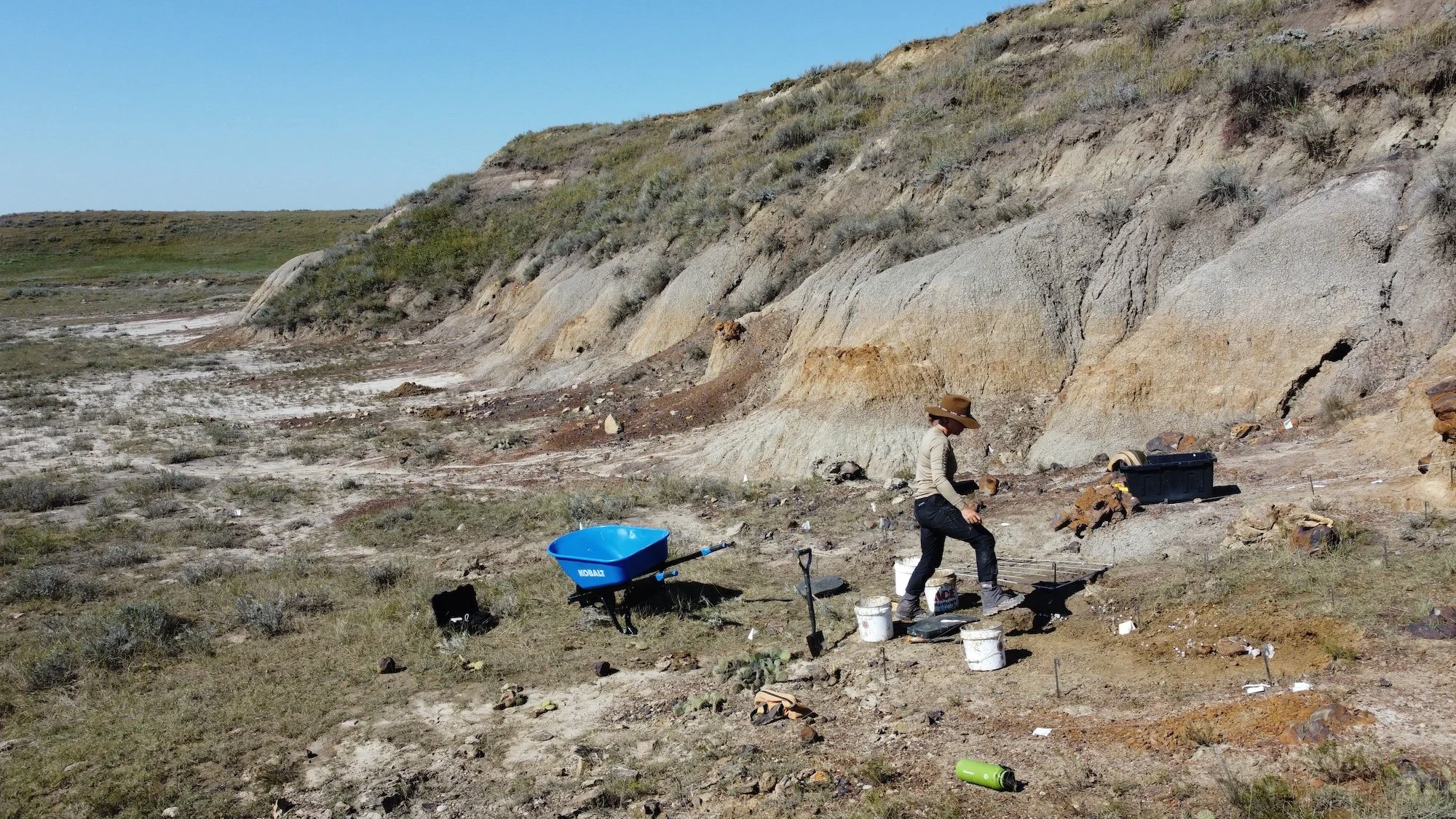 A person wearing a large hat, long-sleeve shirt, and pants, standing in a desert landscape with bare ground, rocks, and sparse vegetation, working with tools near a black container and a wheelbarrow.
