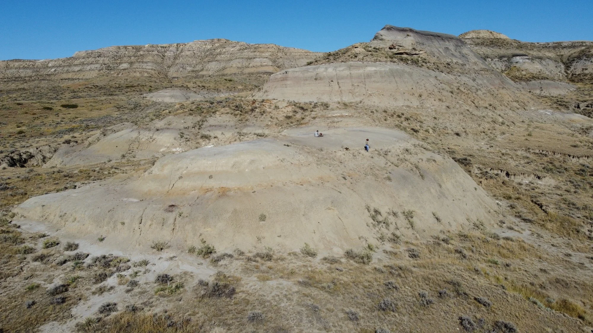 Aerial view of a rocky, desert landscape with sparse vegetation, two people sitting on the ground, and a clear blue sky.
