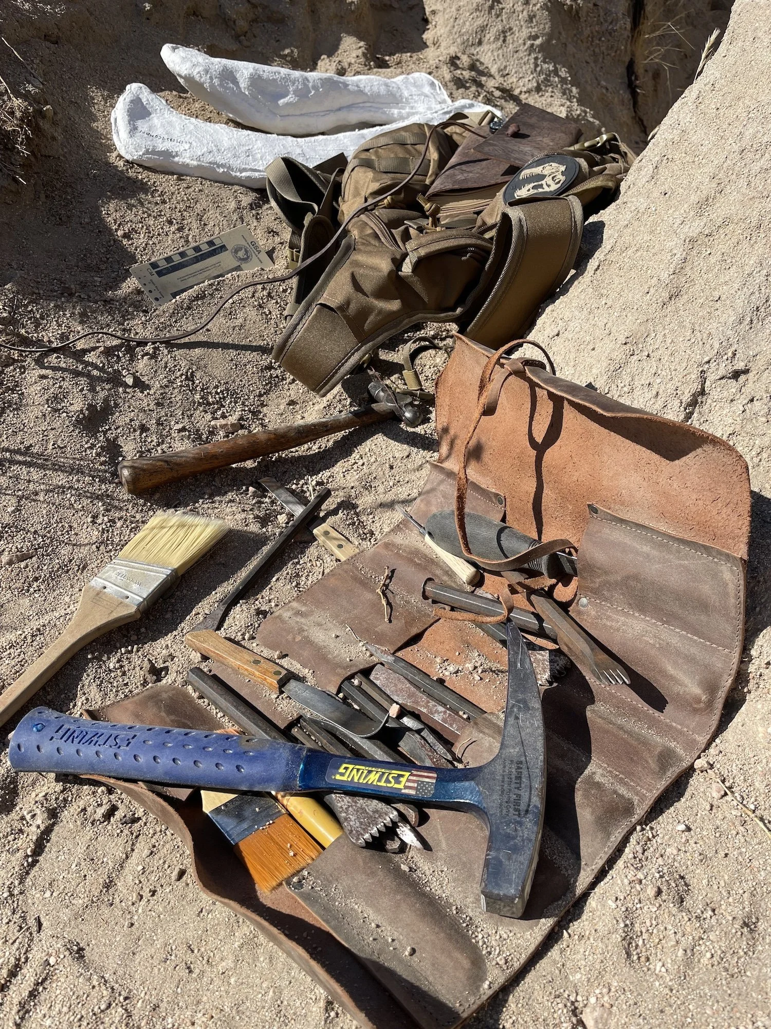 A collection of tools for outdoor work, including a hammer, paintbrush, small saw, and a set of pliers, along with a brown leather toolbox, scattered on sandy desert ground next to a green tactical backpack.
