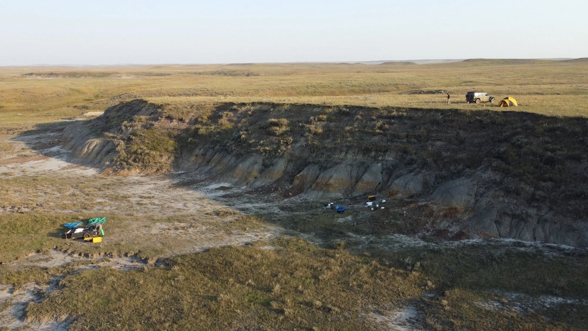 A campsite on a grassy prairie near a steep eroded hillside with cars, tents, and camping supplies.
