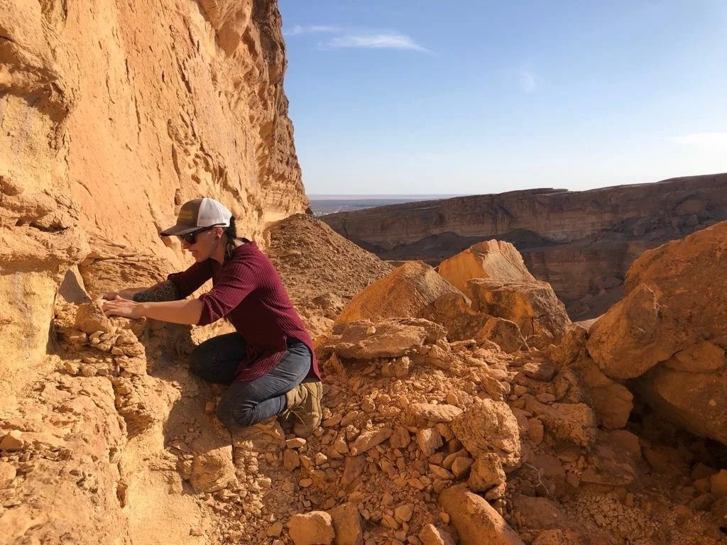 A person crouching on a rocky, desert landscape next to a large cliff, examining or collecting rocks.